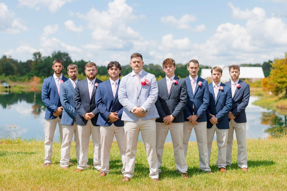 A wedding party strikes playful poses in a line wearing navy suits and gray pants in a scenic outdoor setting.