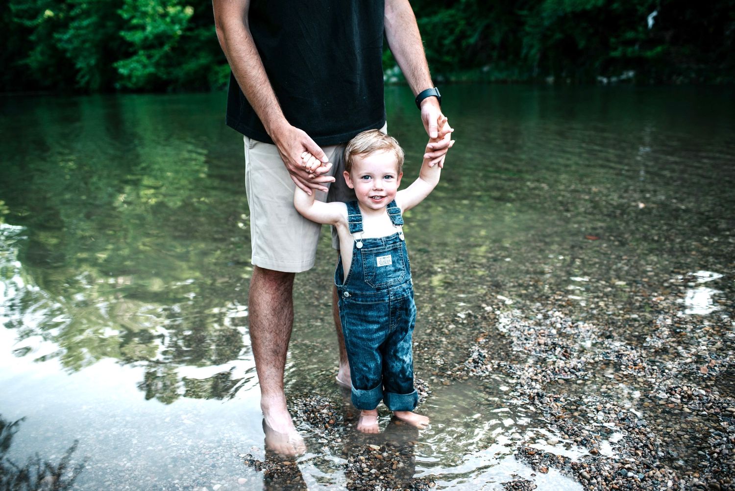 A family enjoys a summer day splashing and playing together in a shallow creek during creek mini sessions with trees in the background.