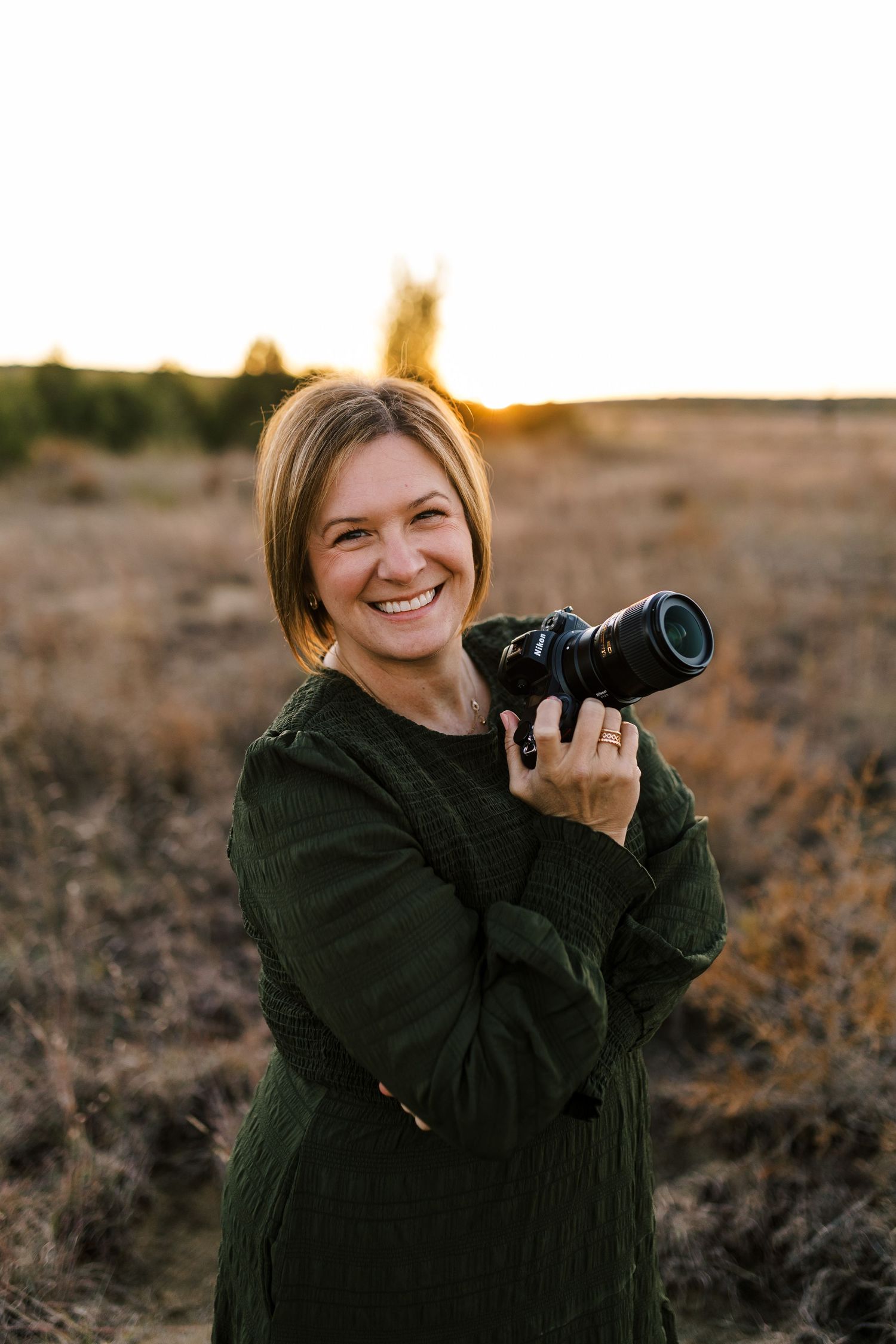 Smiling female photographer holding camera outdoors in golden field at sunset wearing green dress.