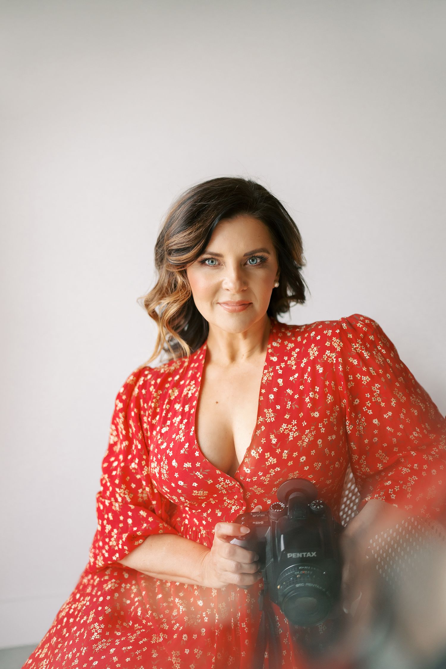 Professional photographer in flowing red polka dot dress holding a camera against light gray background.