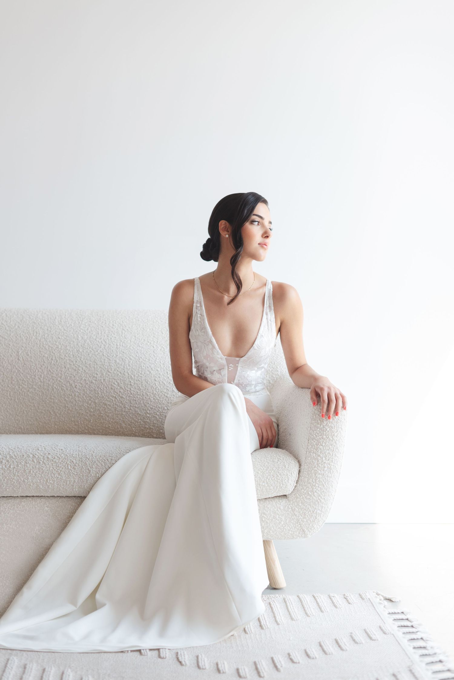 Elegant figure in flowing white wedding gown sits gracefully on cream colored sofa against minimal white backdrop.