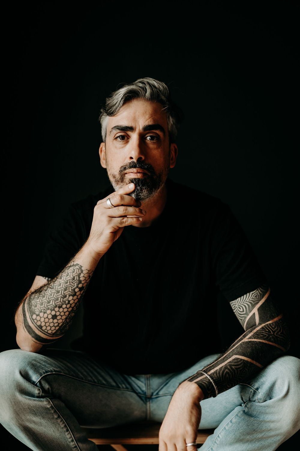 Portrait of person in black shirt with tattoos sitting cross-legged against dark background with contemplative pose.