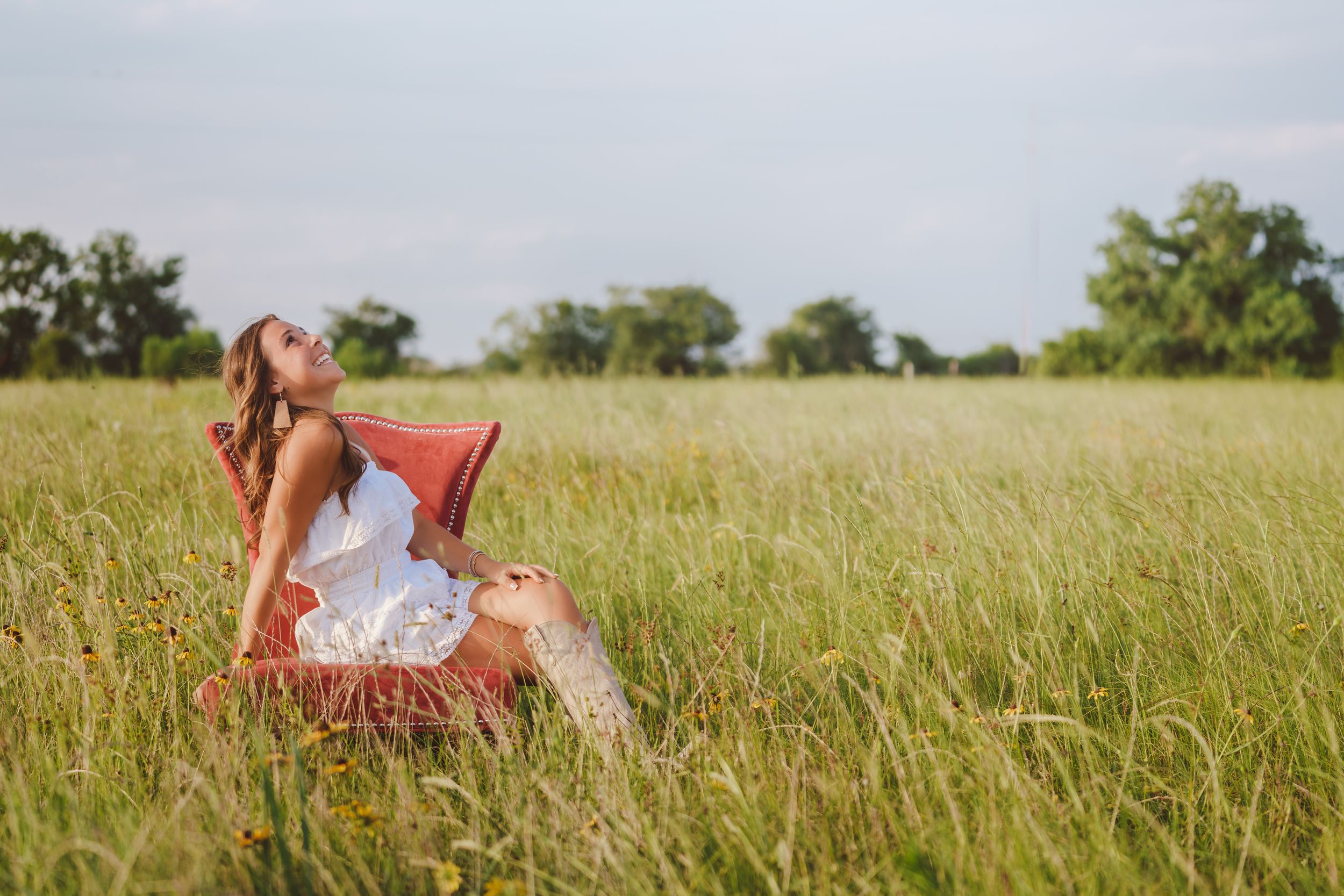 Someone in a white summer dress sits peacefully in a red chair amid tall grass in a sunny meadow.
