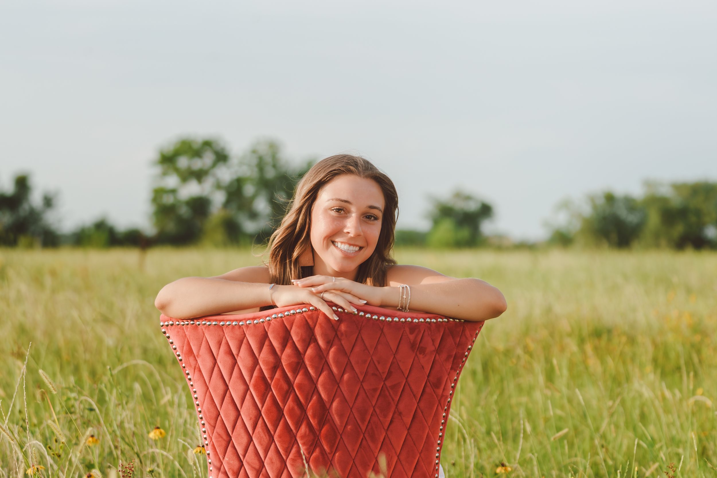 Someone poses cheerfully while leaning on a red woven chair in a sunny summer meadow with tall grass.