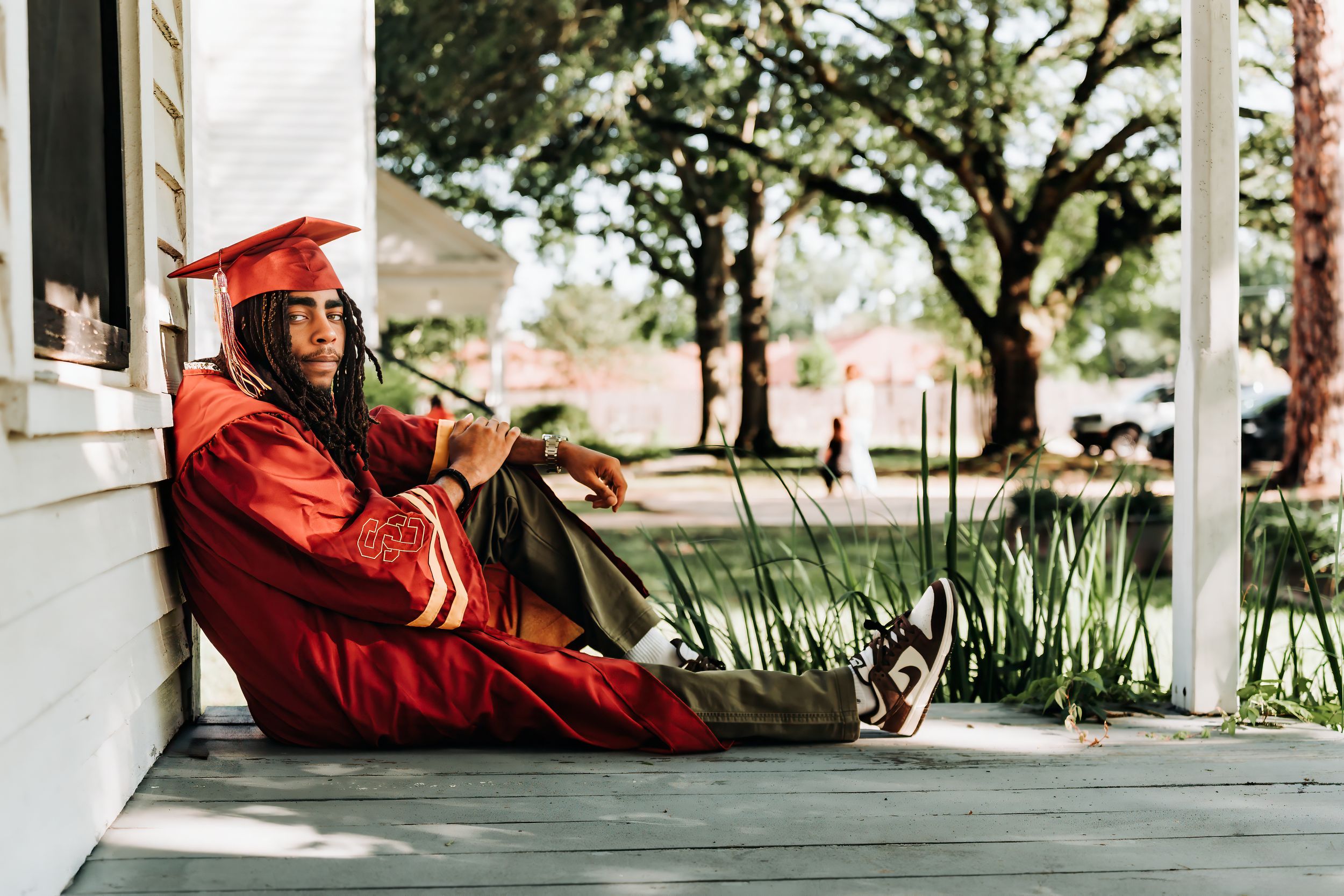 Graduate in red cap and gown sitting relaxed on a porch, celebrating academic achievement outdoors.