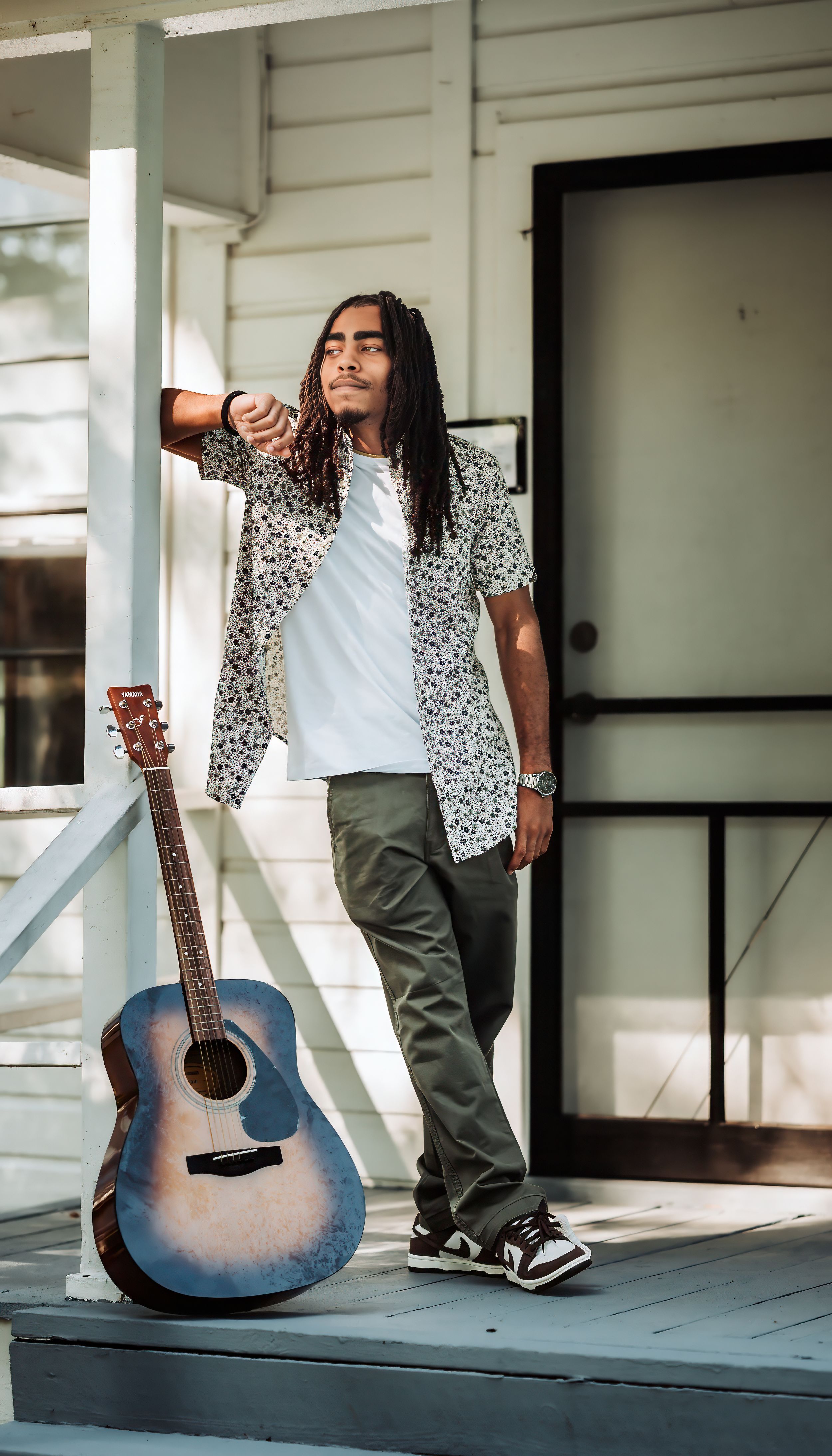 Young man with dreadlocks in floral shirt leaning on porch post with acoustic guitar.