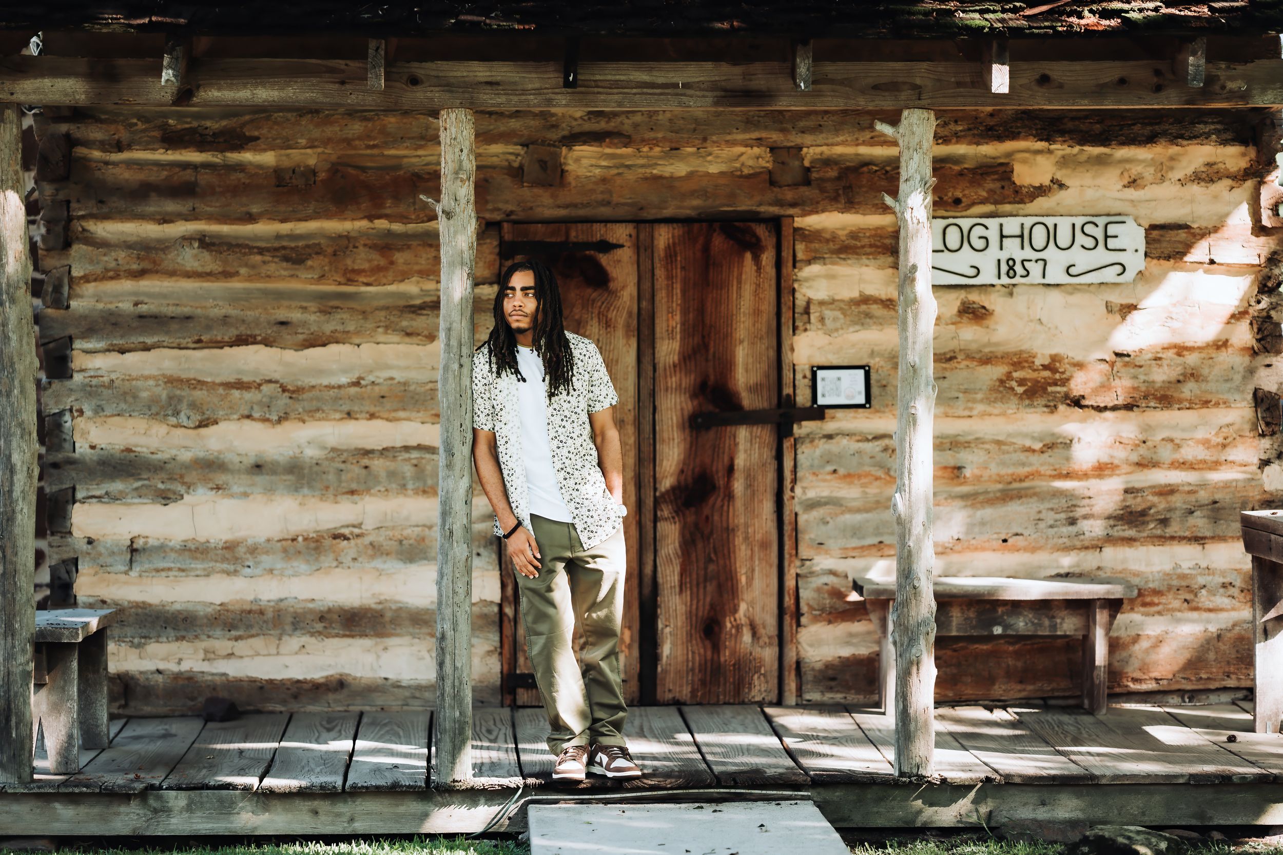 Person with long hair standing on porch of rustic 1857 log cabin Dog House building in natural setting.