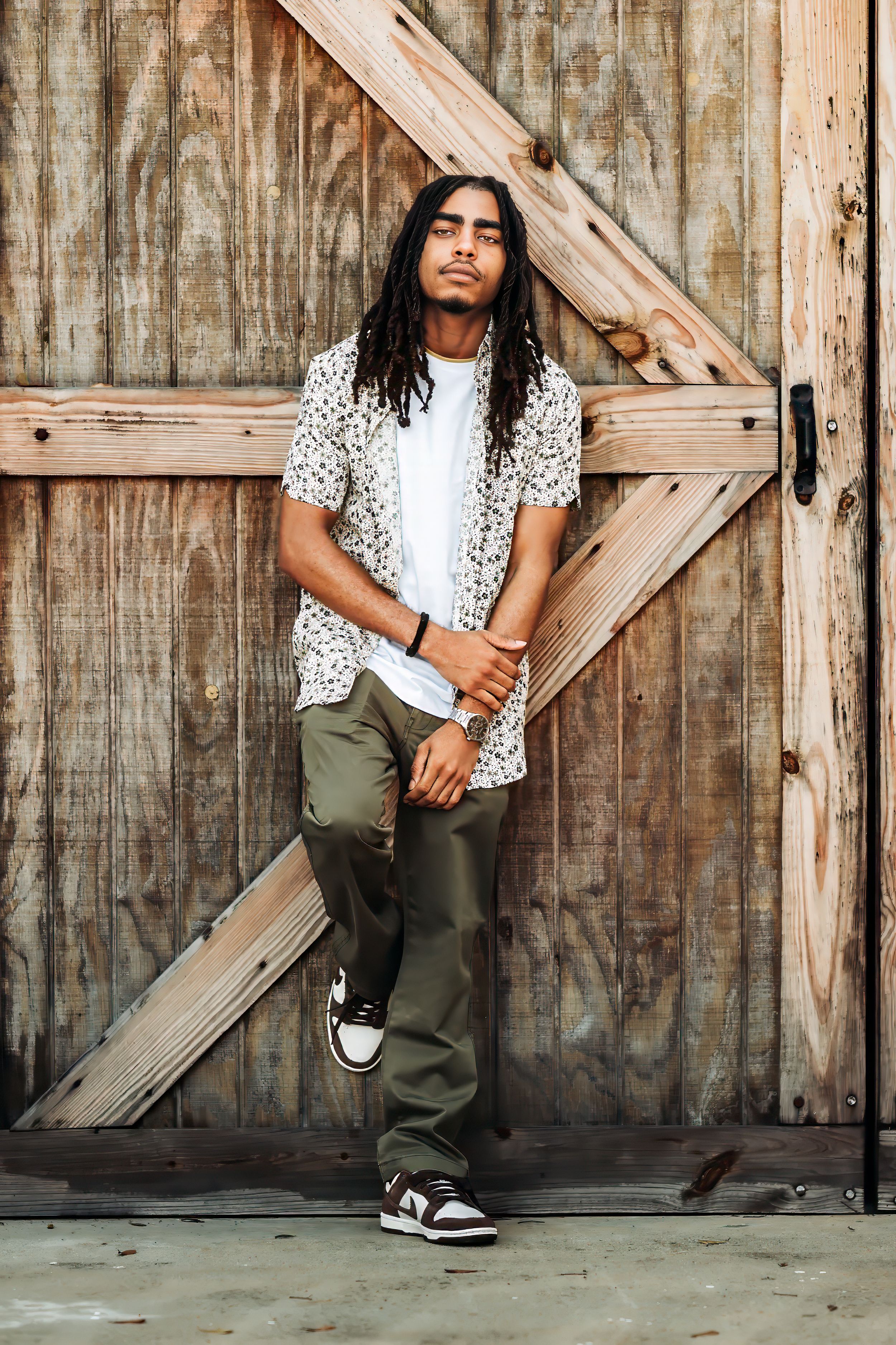 Young man with dreadlocks in floral shirt and olive pants poses confidently against rustic wooden barn door.