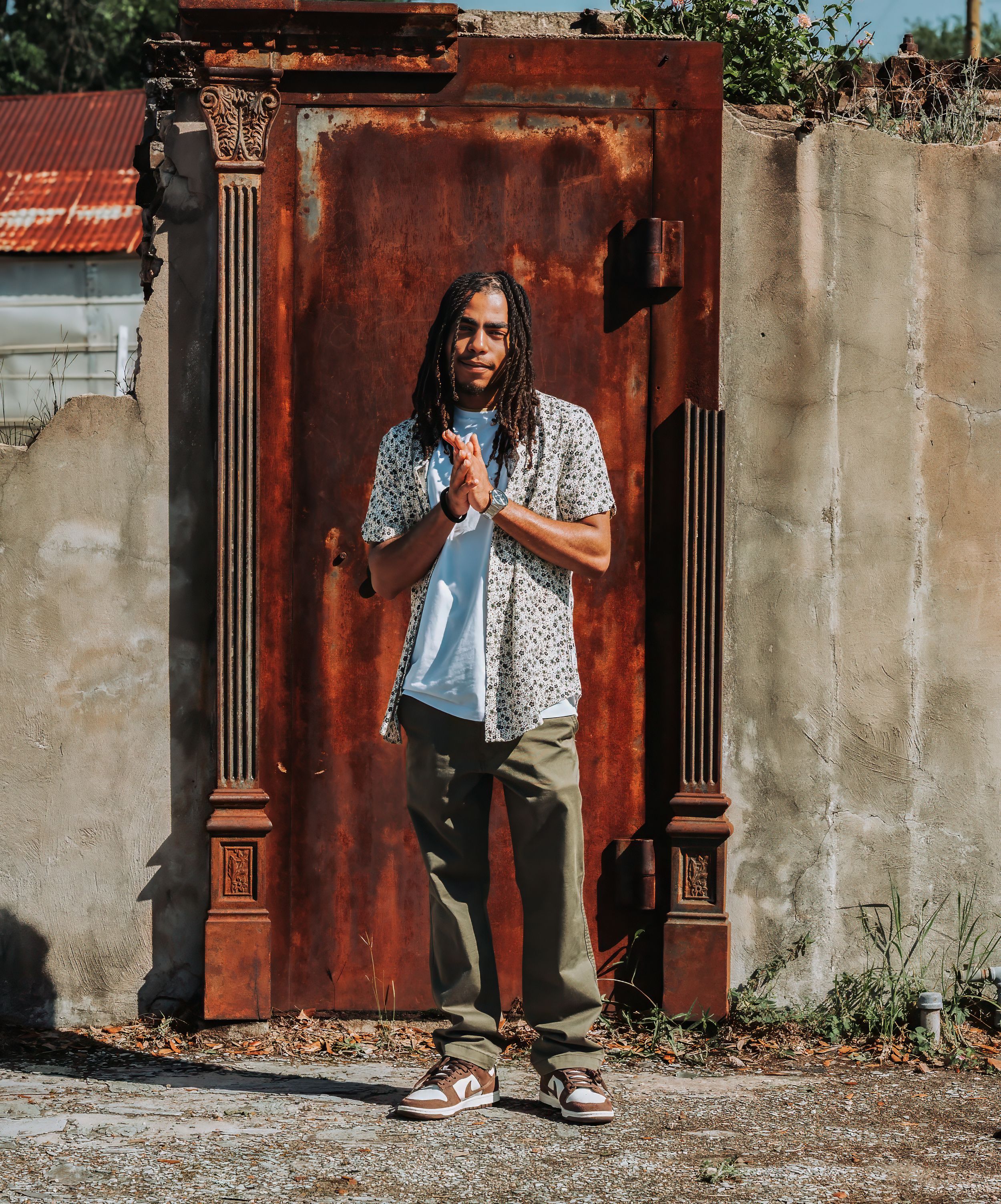 Young man with dreadlocks in floral shirt posing hands clasped before rustic metal door.