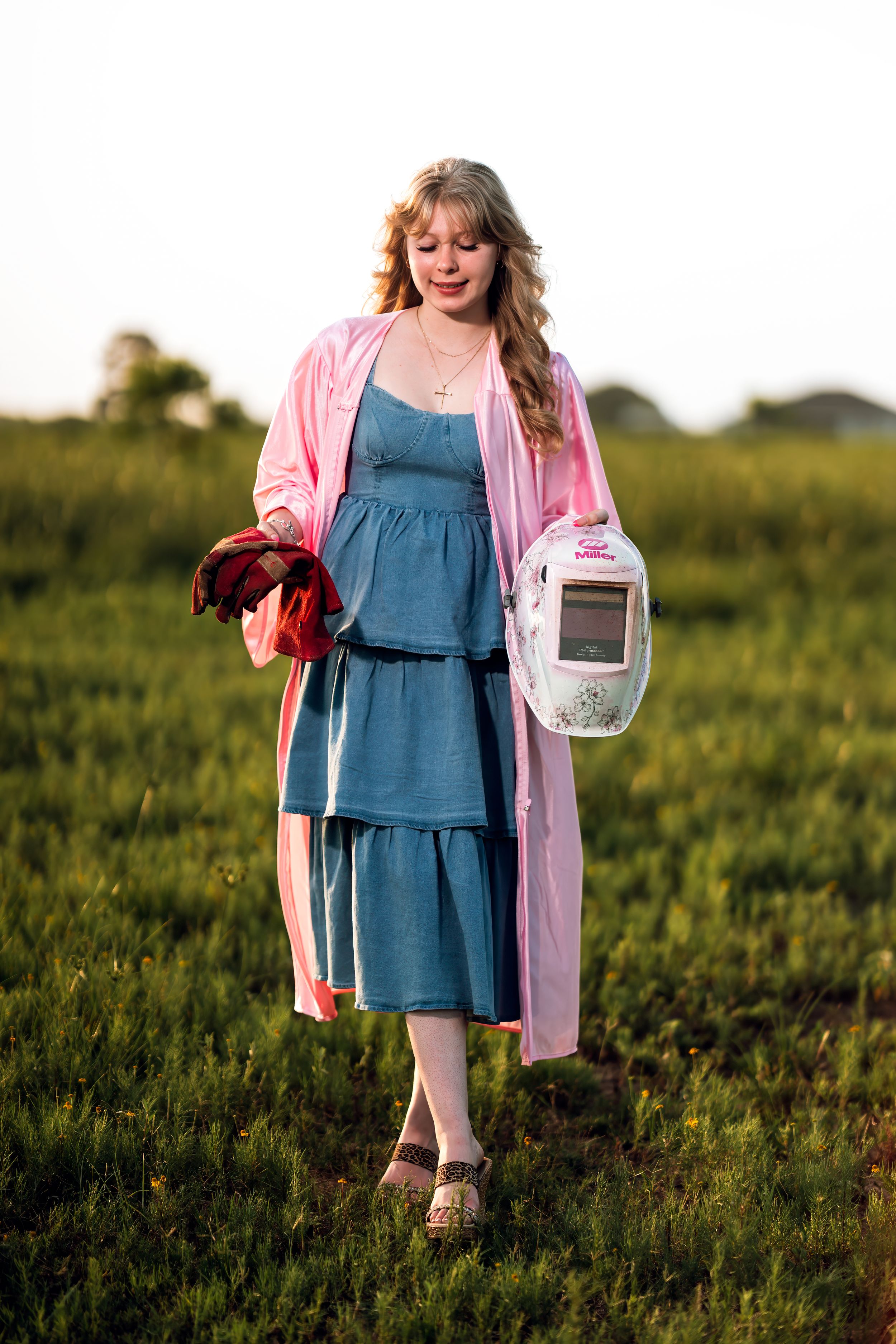Young woman in pink cardigan and blue dress holding a welding mask and gloves in a green field.
