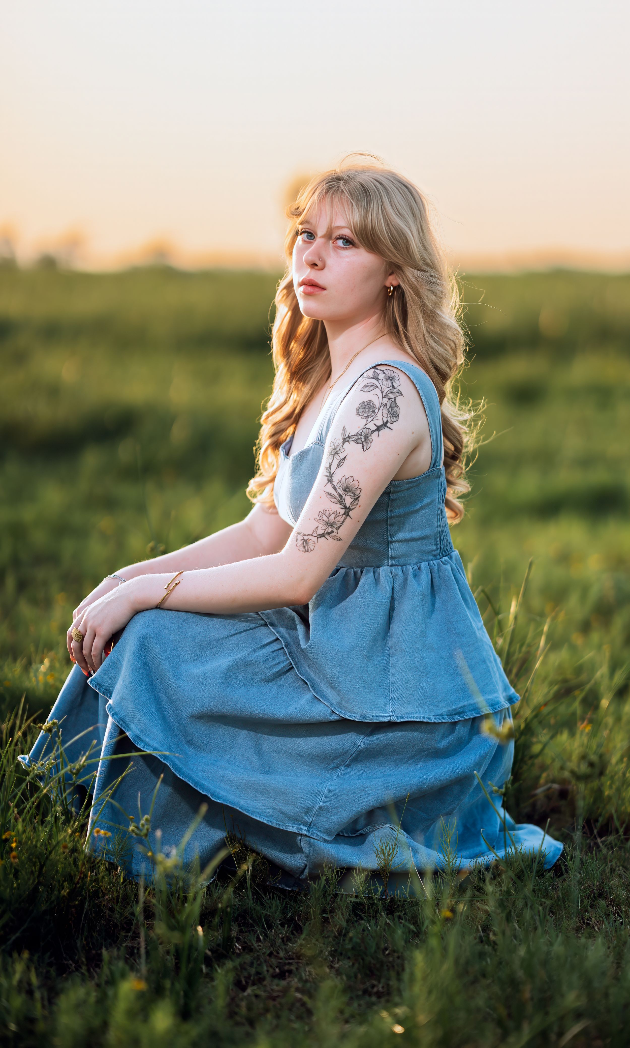 Young blonde woman with floral arm tattoo in blue dress sitting in green field during golden hour sunset.