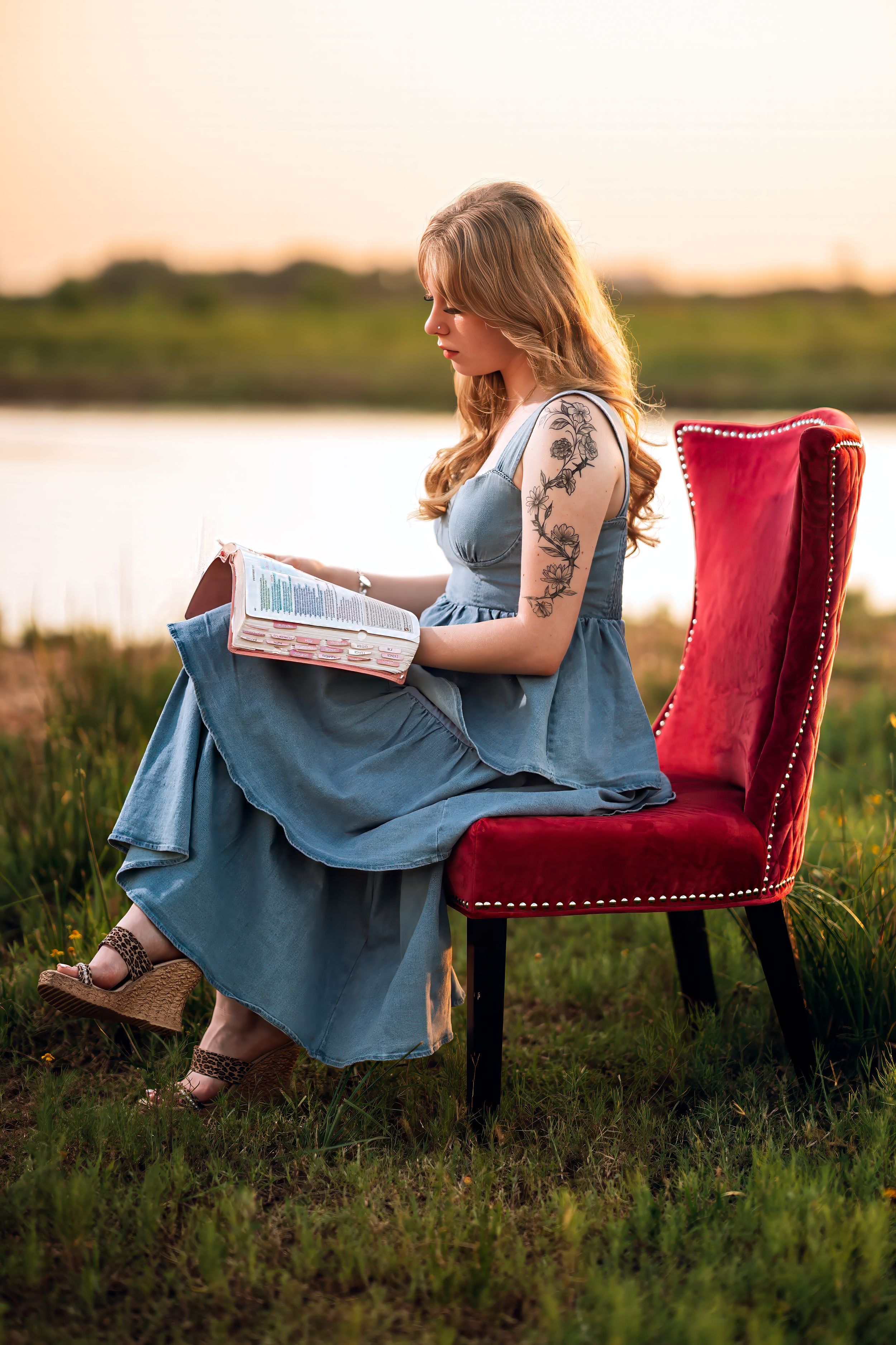 Woman with tattoos in blue dress reads book on red velvet chair outdoors at sunset near water.