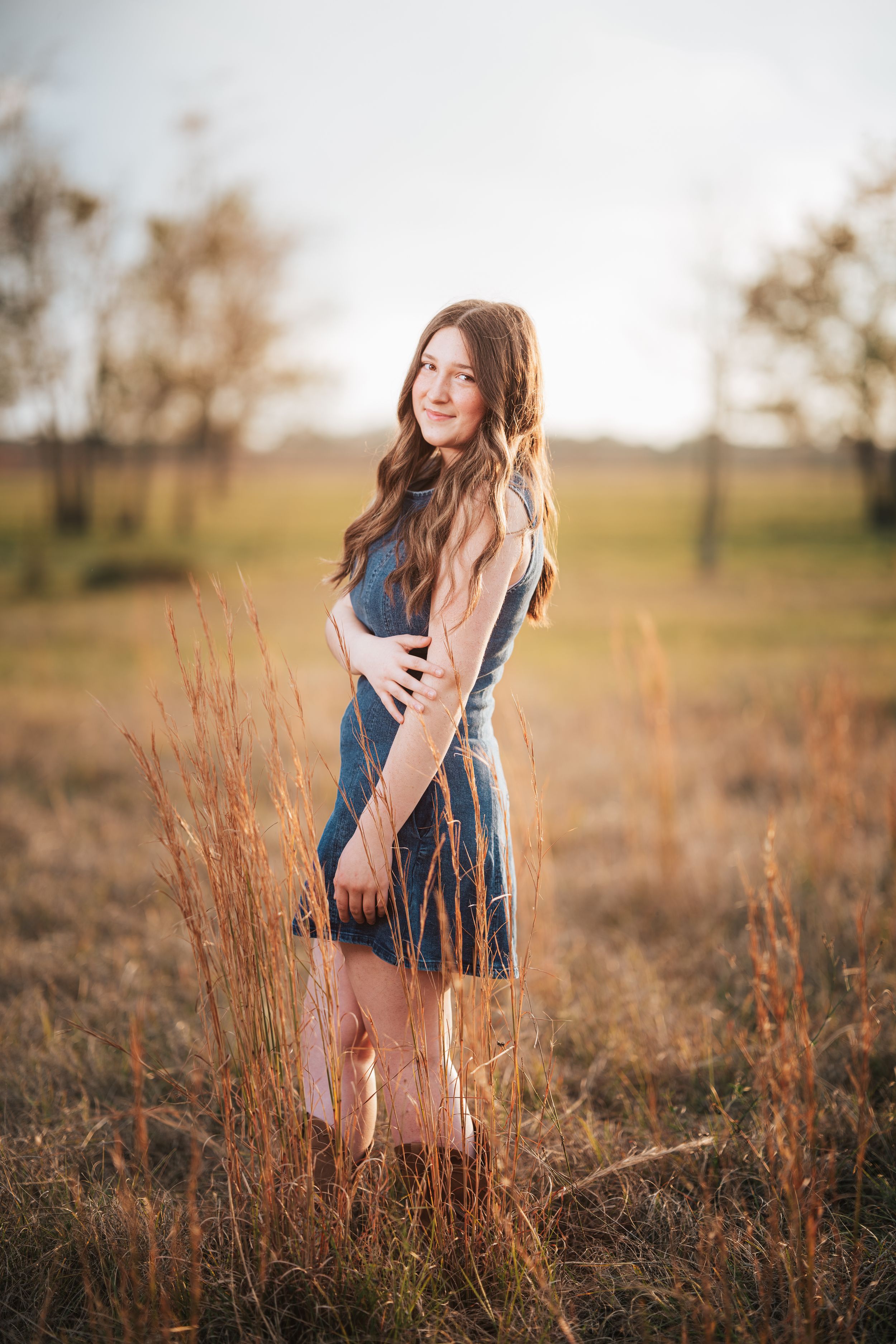 A figure in a denim dress poses gracefully among tall autumn grasses in a sunlit field.