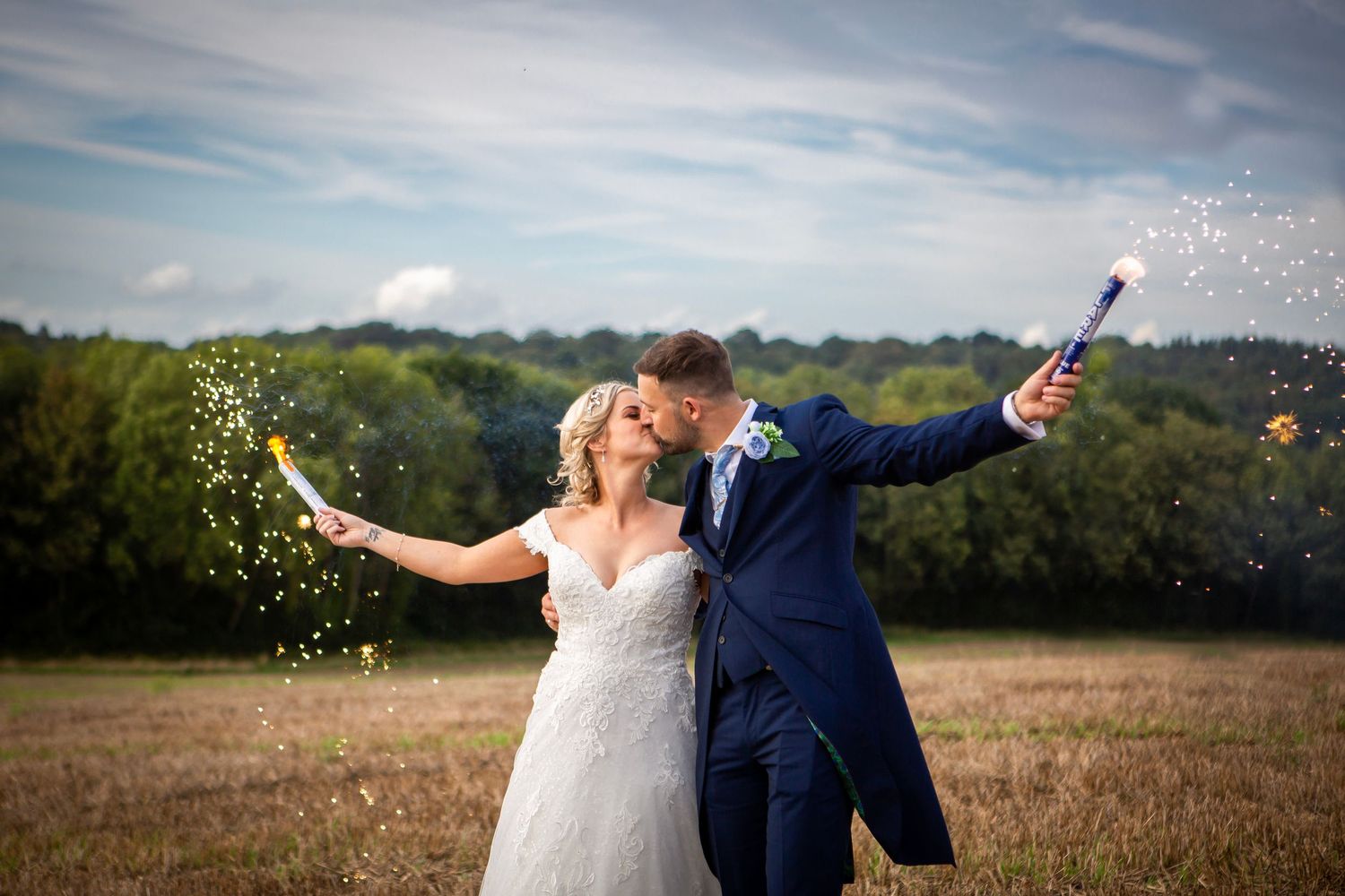 Newlyweds celebrate in a golden field holding sparkling confetti poppers while sharing a romantic kiss outdoors.