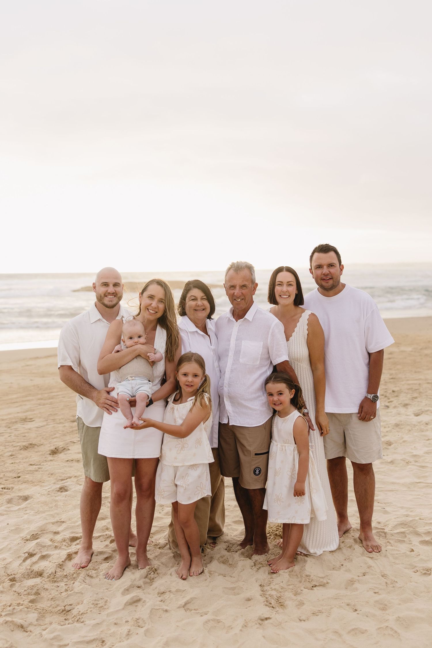 Multi-generational family wearing coordinated white and beige outfits poses together on a beach at sunset.