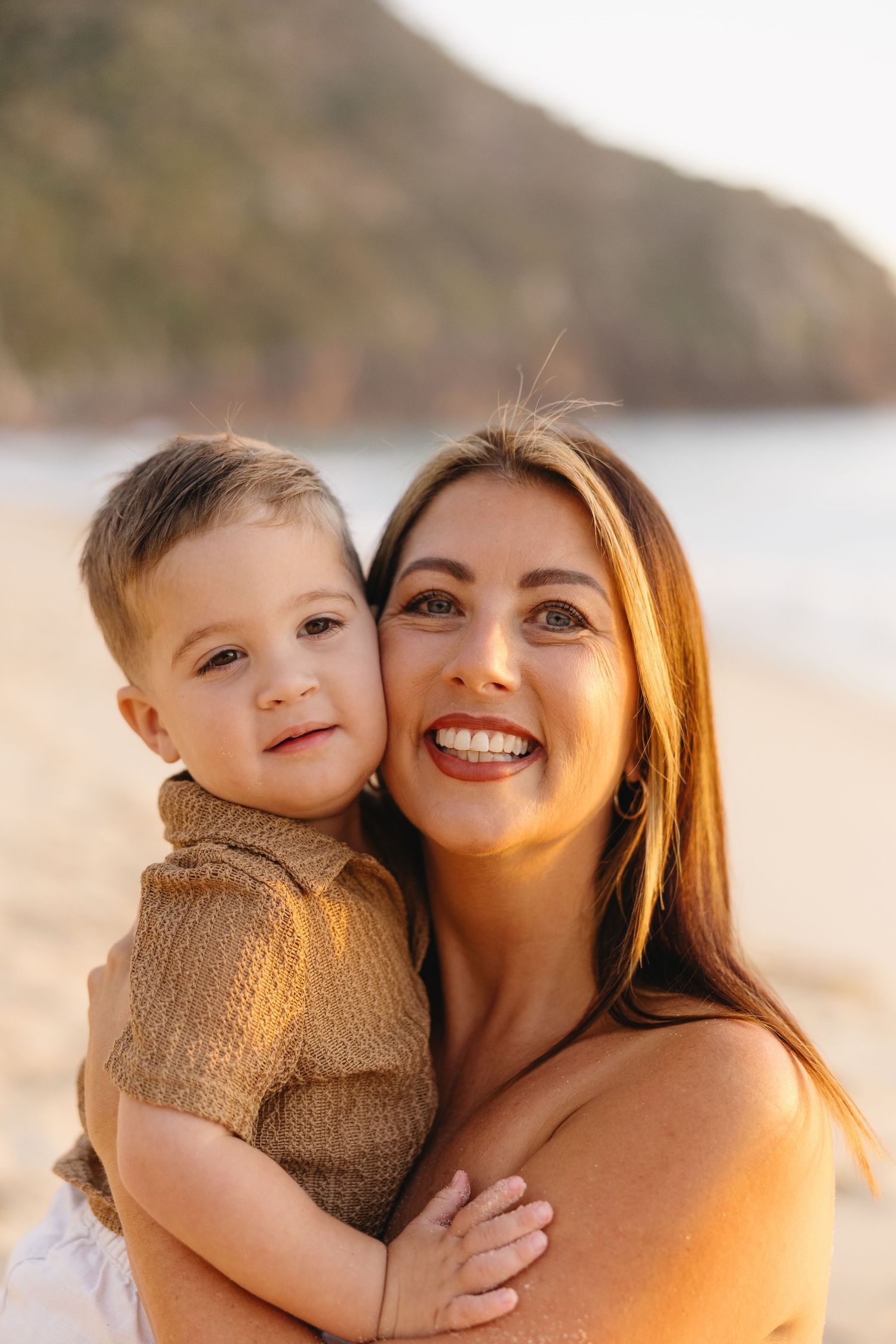 Parent and child share a tender moment at sunset on a beach with golden lighting and coastal cliffs in background.