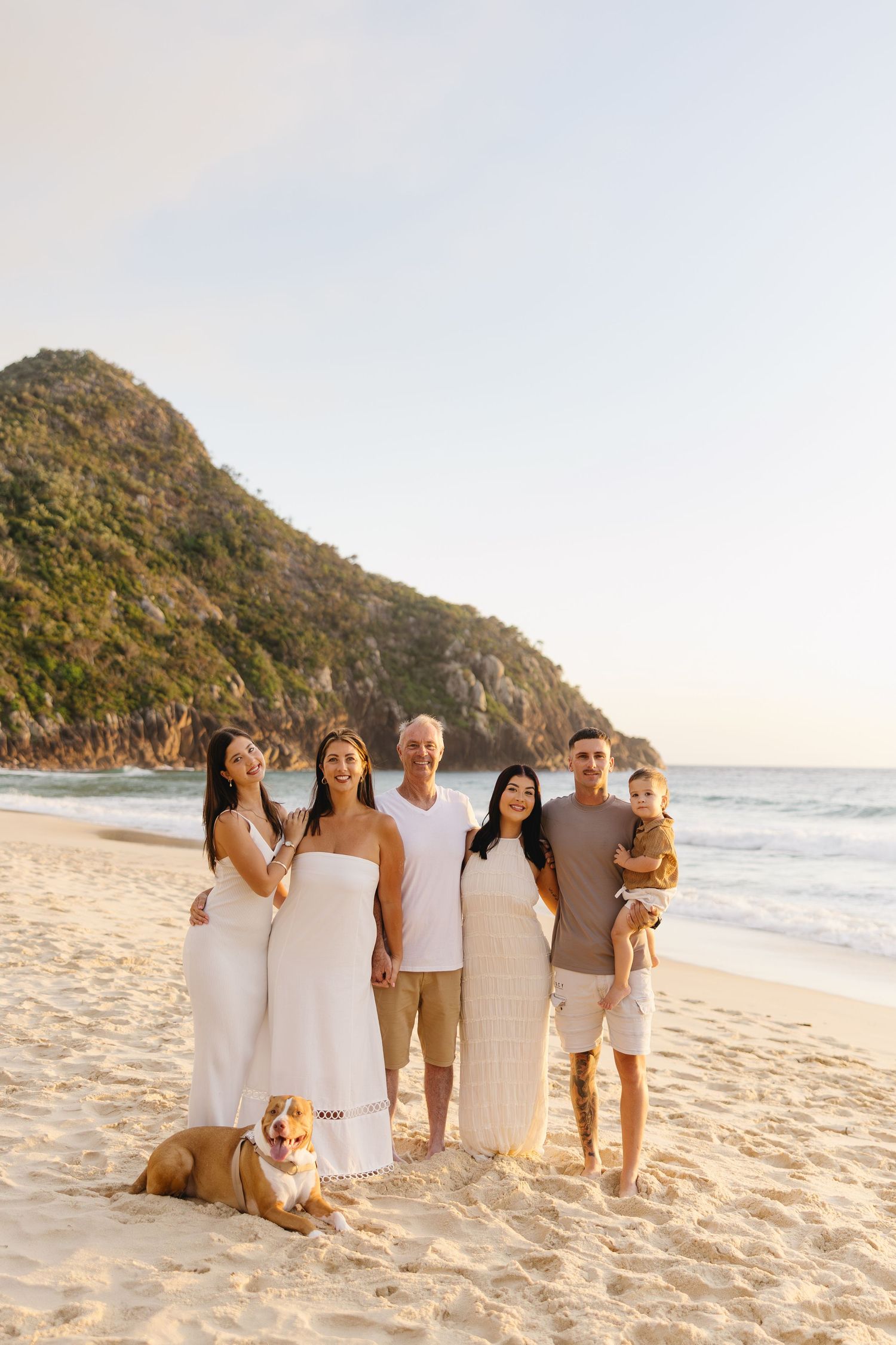 A group in white and beige outfits poses together with their dog on a scenic beach at sunset with mountains in background.