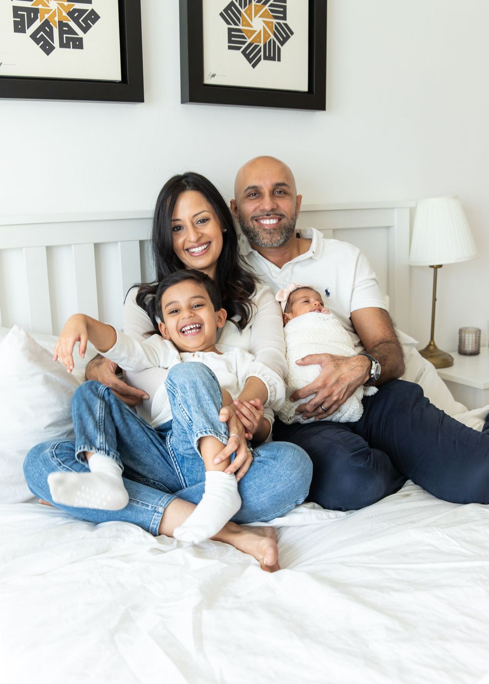 Portrait of family sat on their bed at-home laughing while cuddling baby in swaddle and toddler sibling in Dubai newborn photoshoot