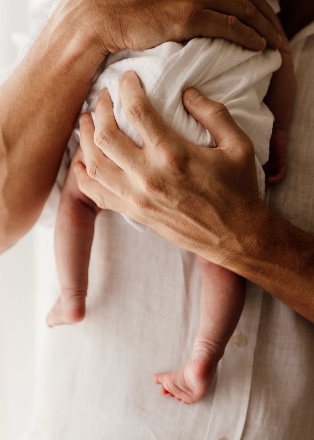 Professional lifestyle newborn photoshoot at-home showing a close up portrait of dad's hand supporting baby - focus on baby's legs and feet