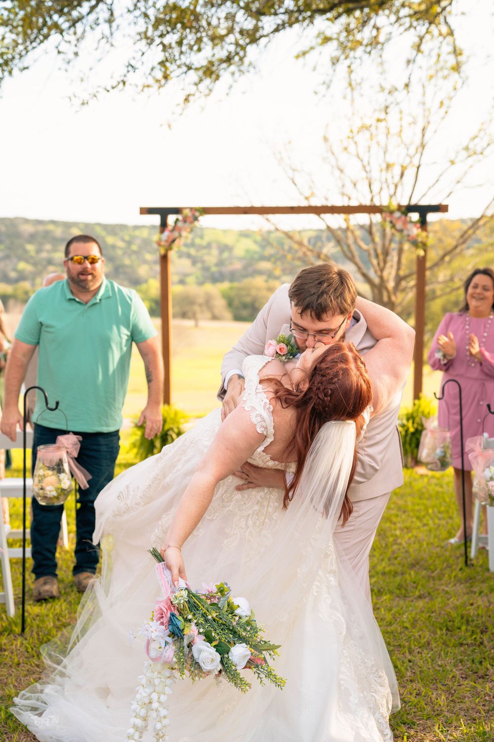 Romantic dip kiss during outdoor wedding ceremony with floral arch backdrop.