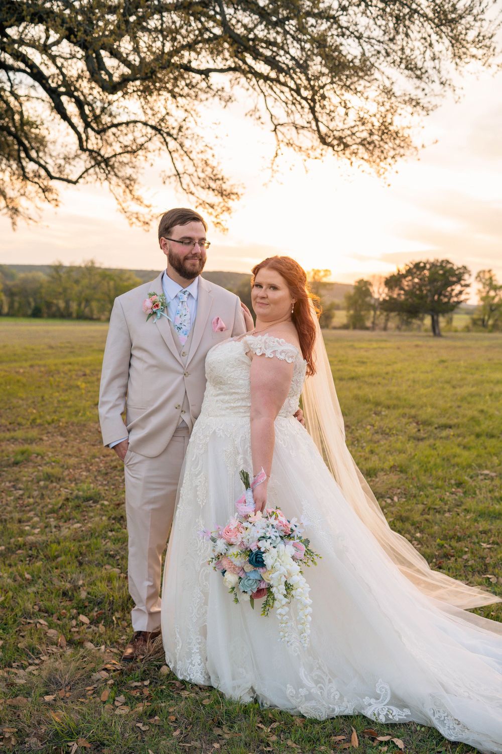 A wedding couple poses in a field during golden hour with a flowing veil and floral bouquet.