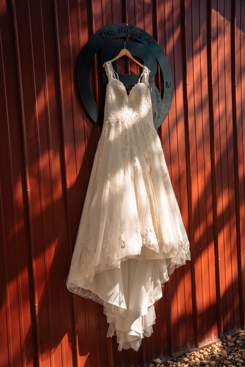 White wedding dress hanging against rustic red barn wall with dramatic shadows.