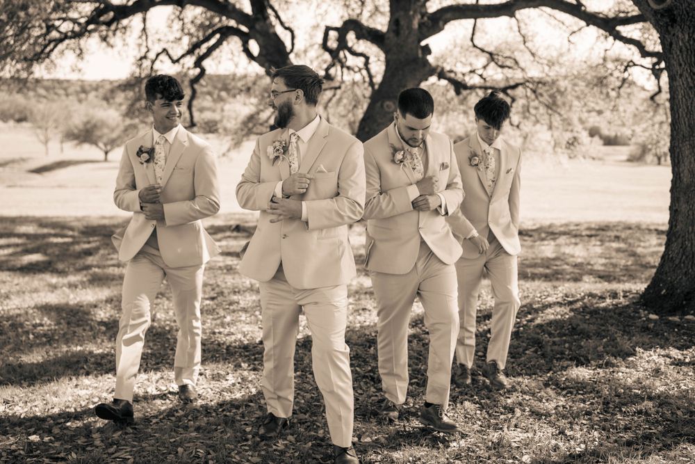 Wedding party in light colored attire poses under tree branches in sepia toned photo.
