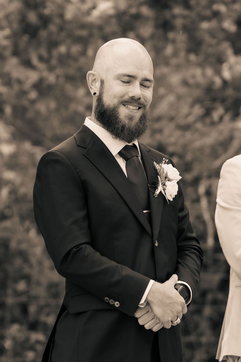 A groom in a black suit and tie smiles during a wedding ceremony with a floral boutonniere.