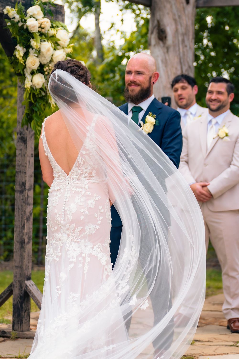A bride in a lace dress with flowing veil joins her wedding party on a wooden deck adorned with floral decorations.