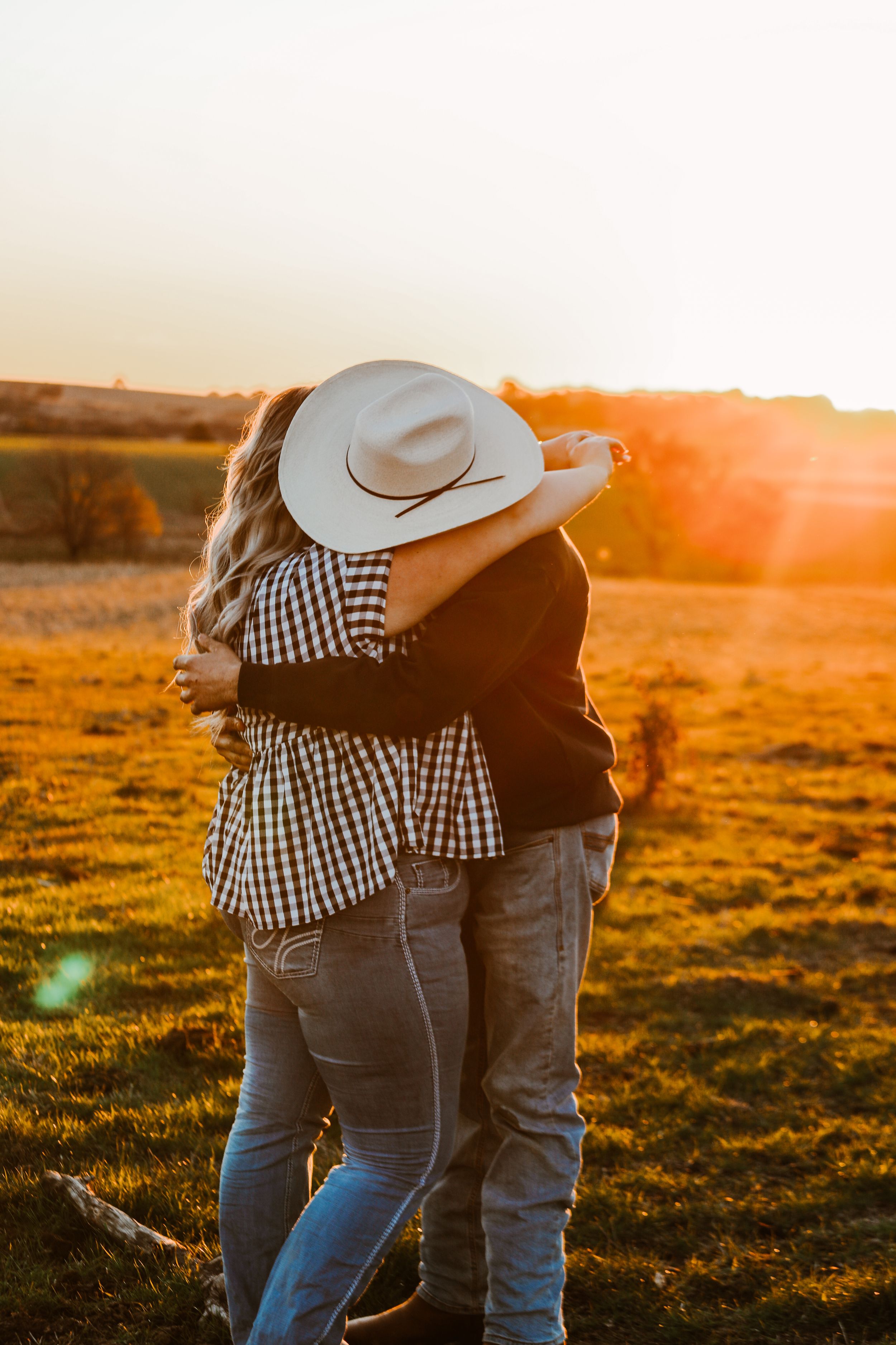 Couple in country attire embracing at sunset with cowboy hat silhouette.