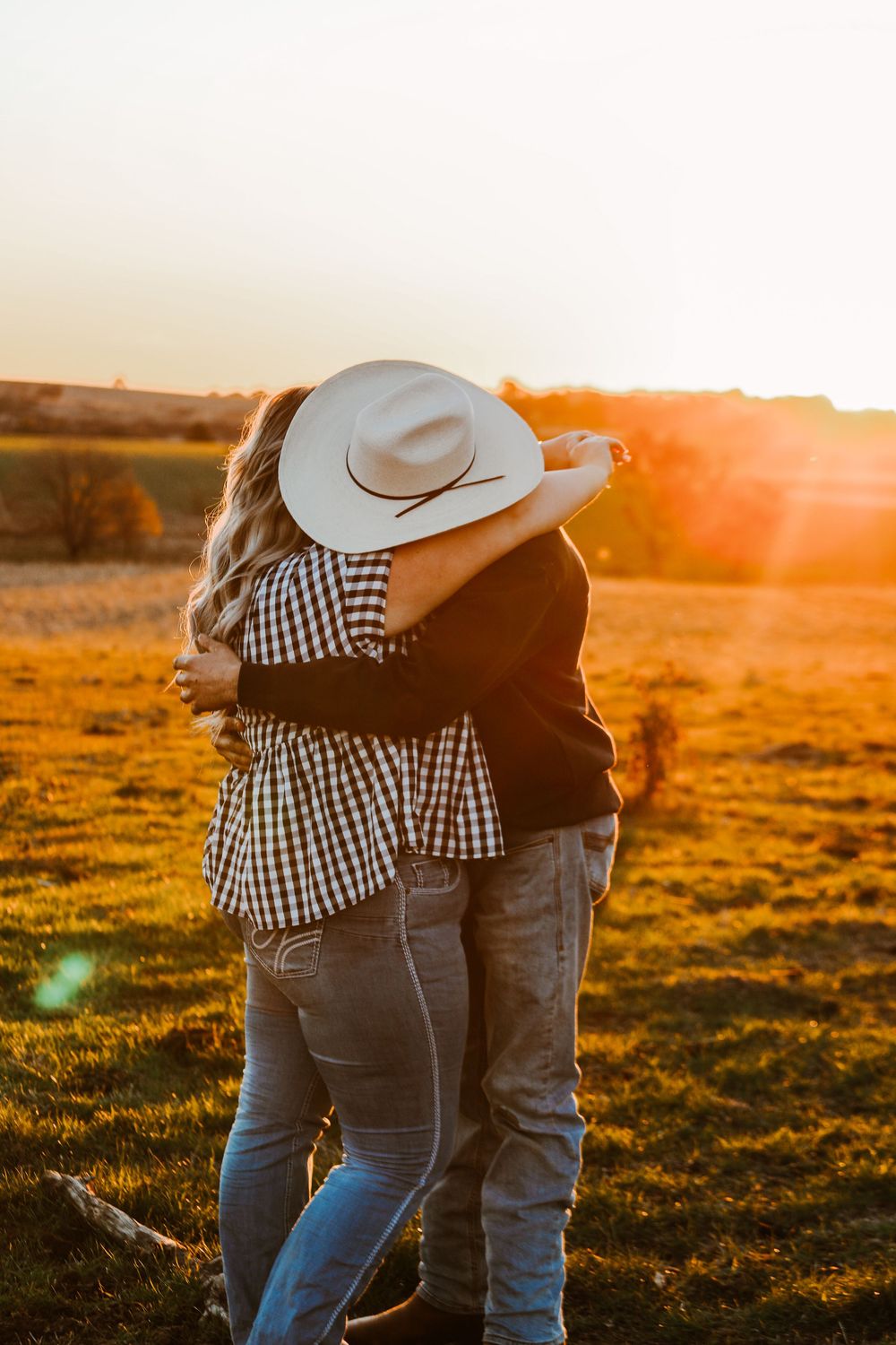 Couple in country attire embracing at sunset with cowboy hat silhouette.
