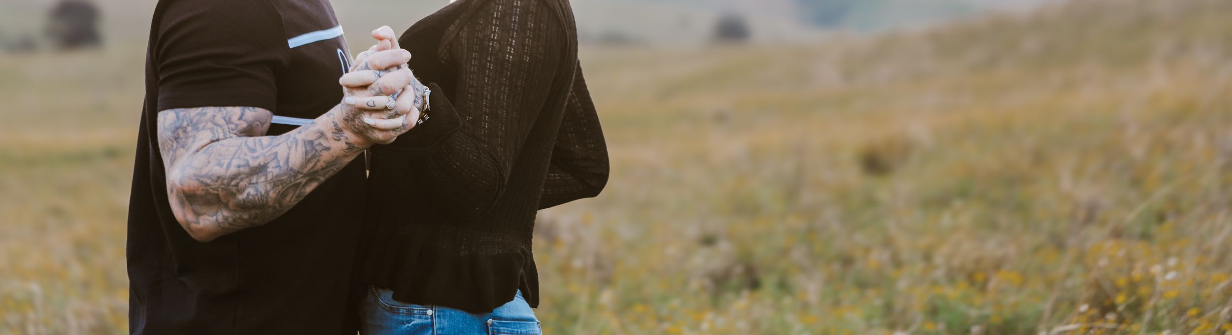 Close-up of clasped hands with tattoos against a rural landscape backdrop.