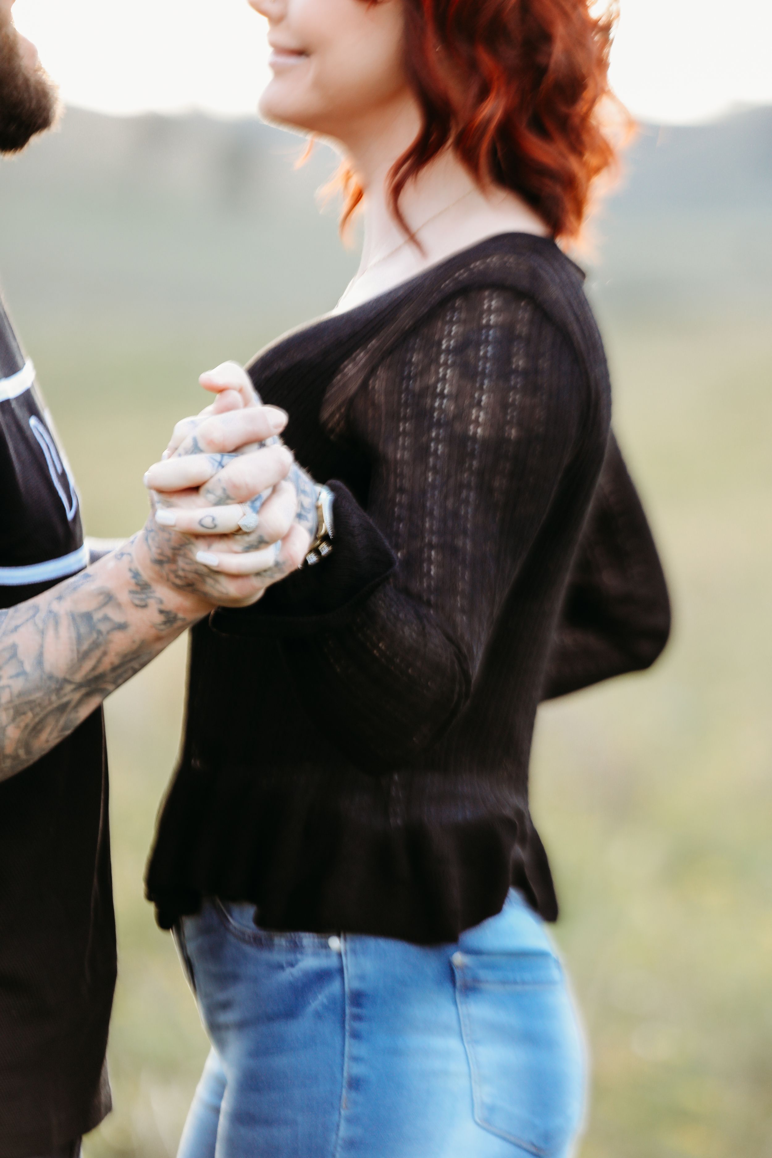 Side view of couple holding hands with one wearing black sweater and jeans in countryside setting.