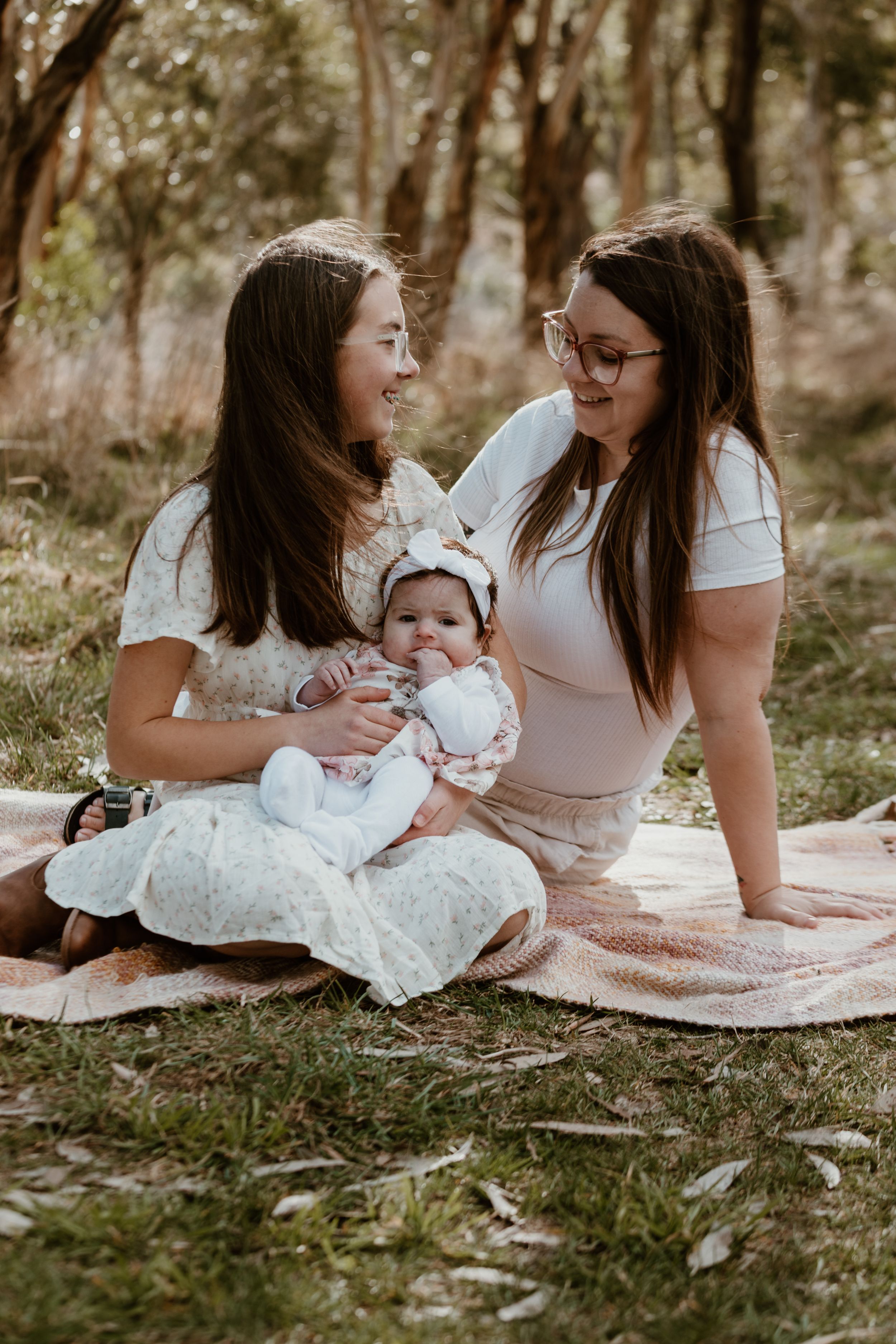 People sitting on blanket outdoors sharing tender moment with baby in white dress.