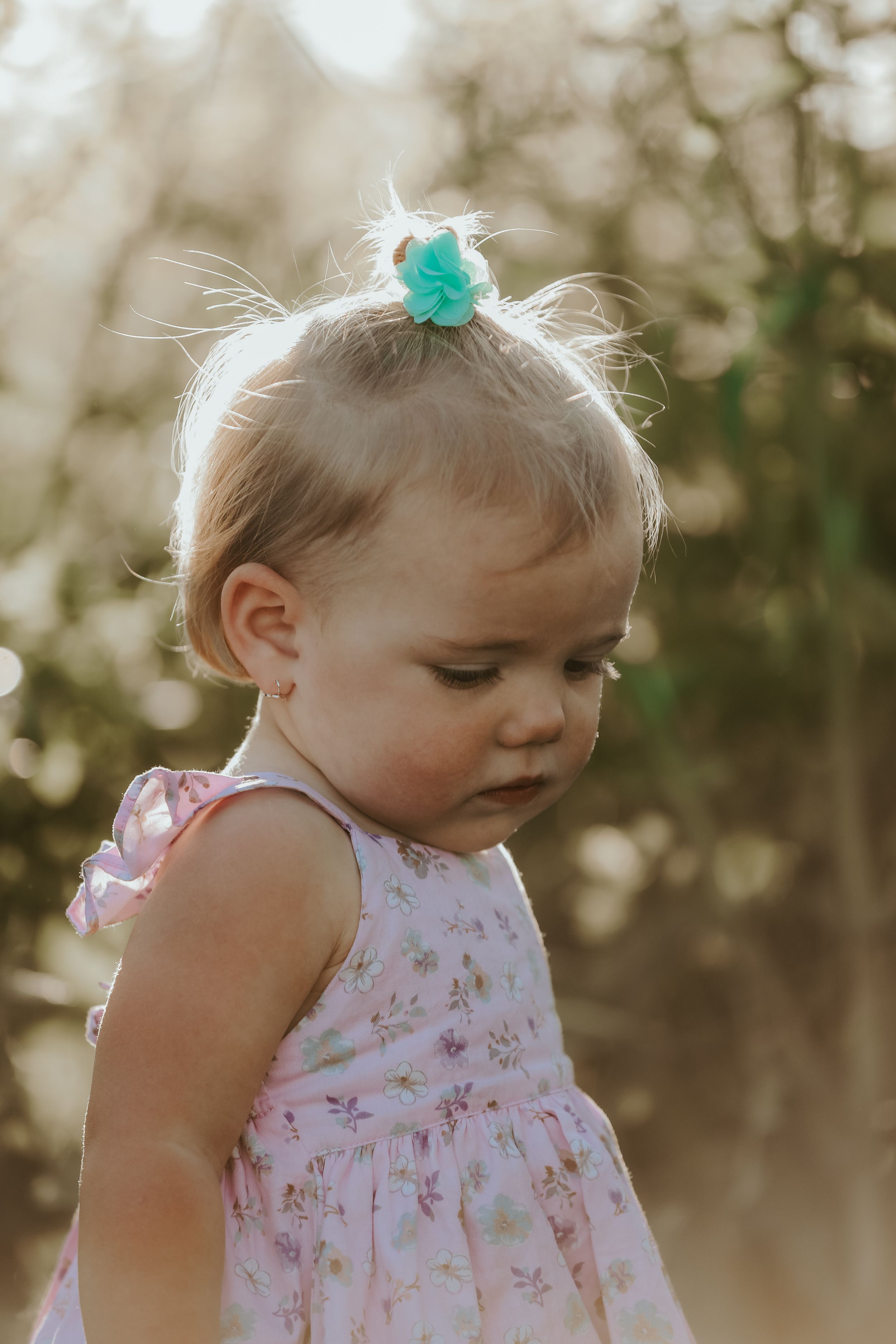 Toddler in floral dress with teal hair bow looking down.