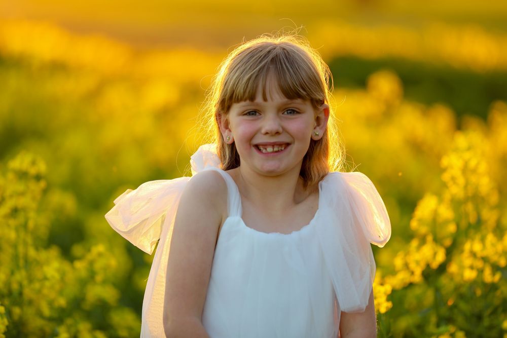 Young child in white dress running through golden canola field at sunset.