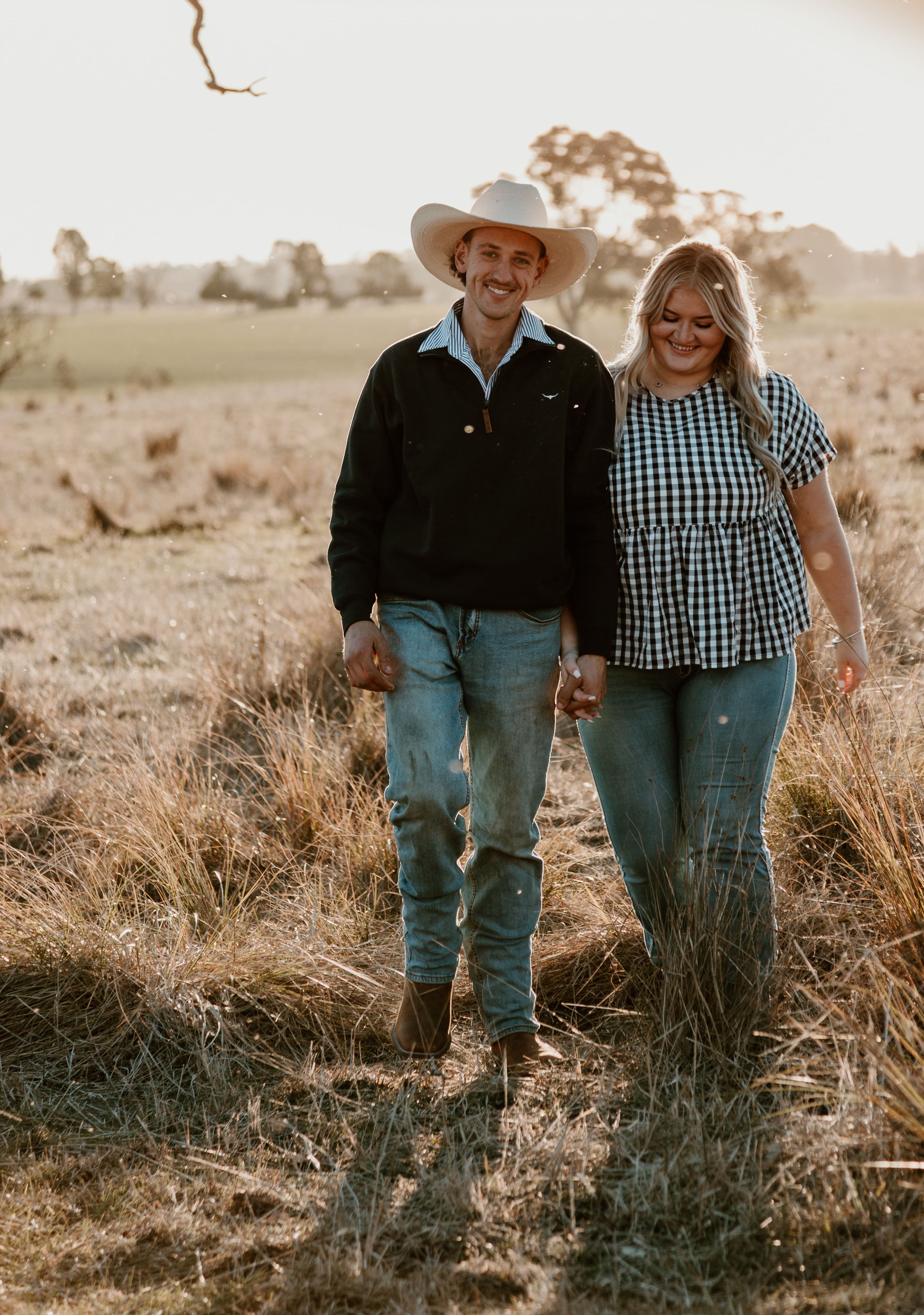 Couple in western attire walking together through rustic countryside at sunset.