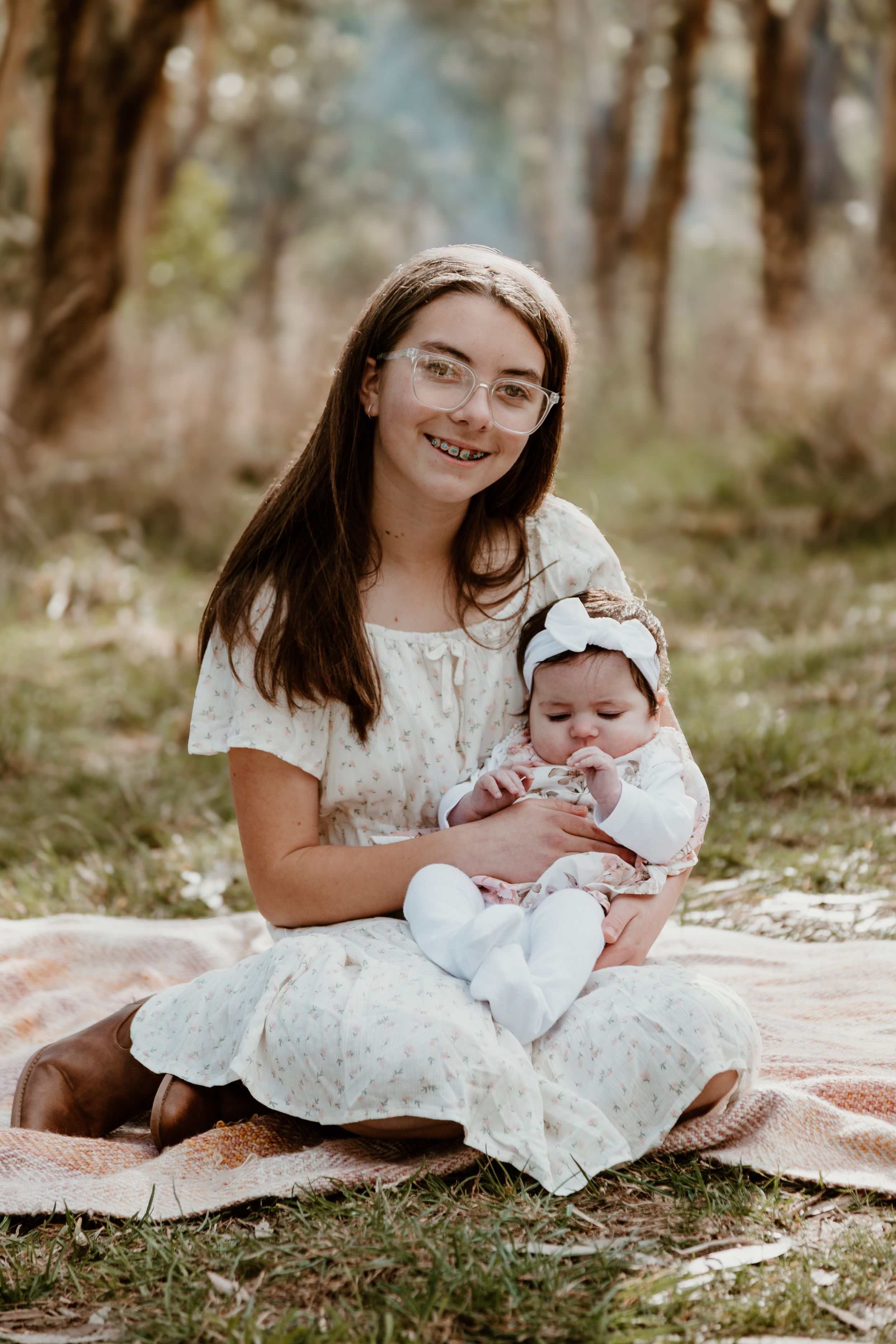 Person in white lace dress holding infant in matching white outfit on grass.
