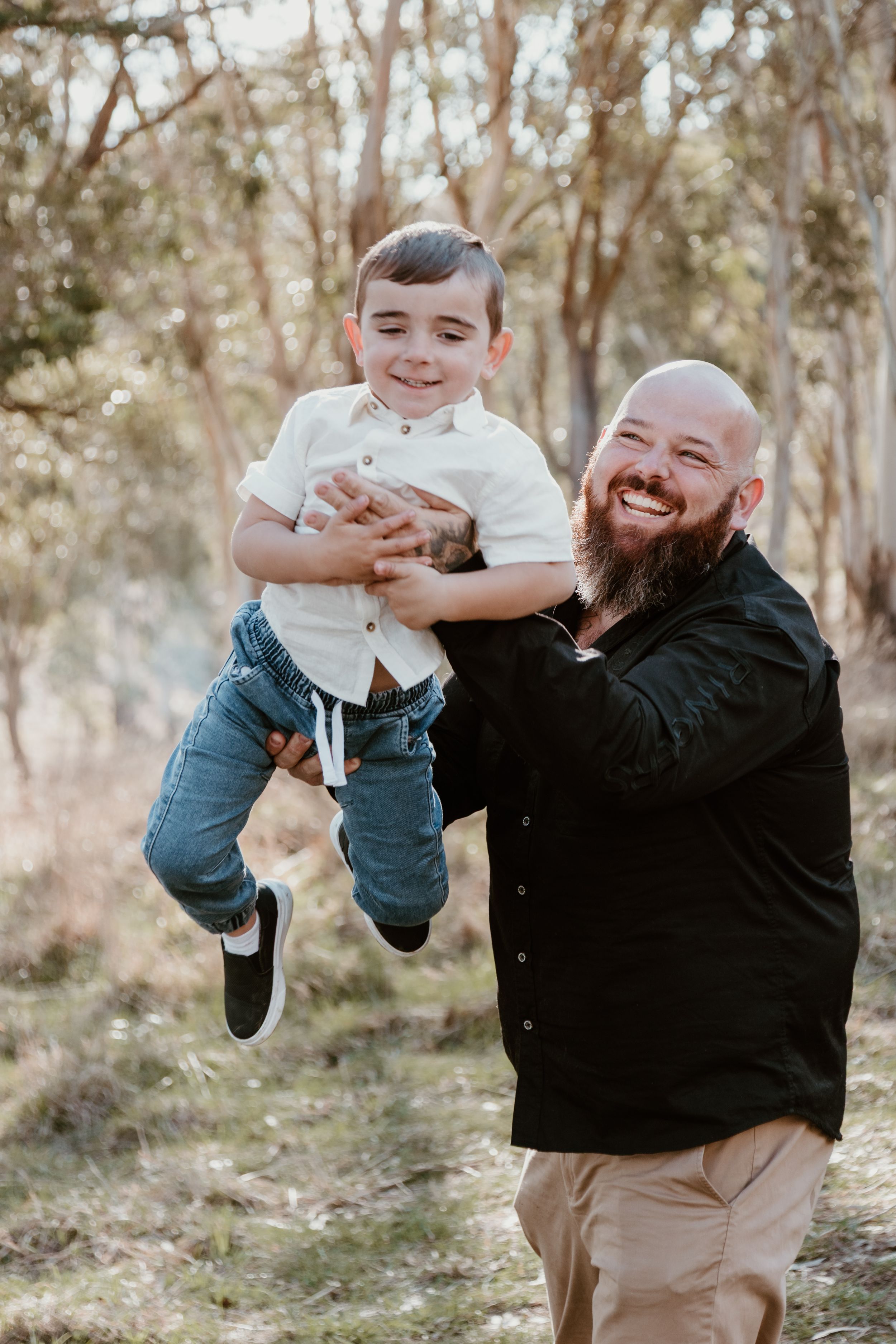 Person with beard playing with baby in white outfit outdoors.
