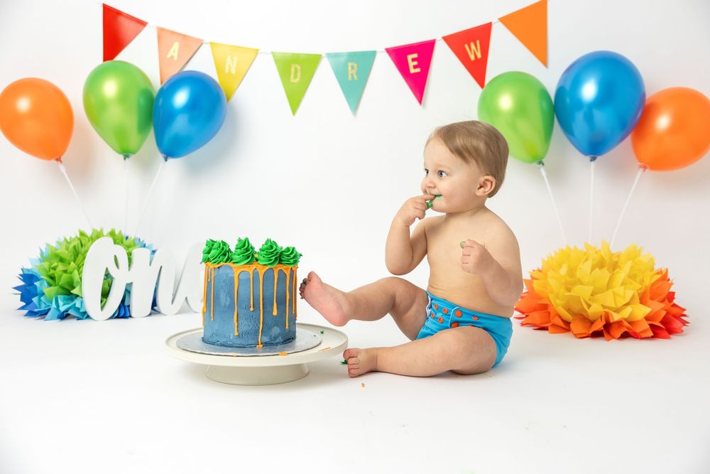 Festive birthday cake smash photo setup with colorful balloons, banners and decorative pom poms for celebration.