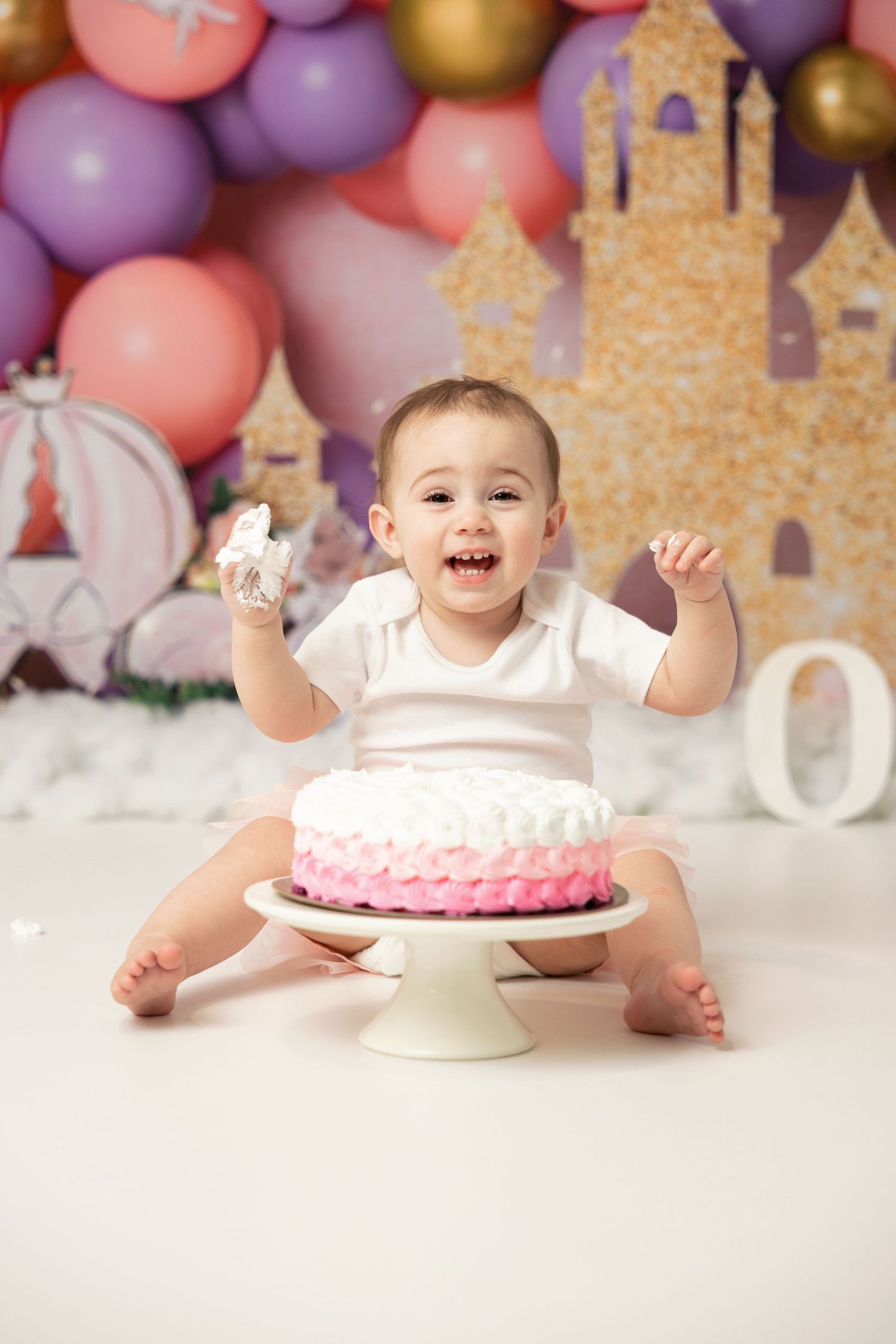 Pink and purple balloon backdrop with cake smash setup featuring a pink ombre cake on white pedestal stand.
