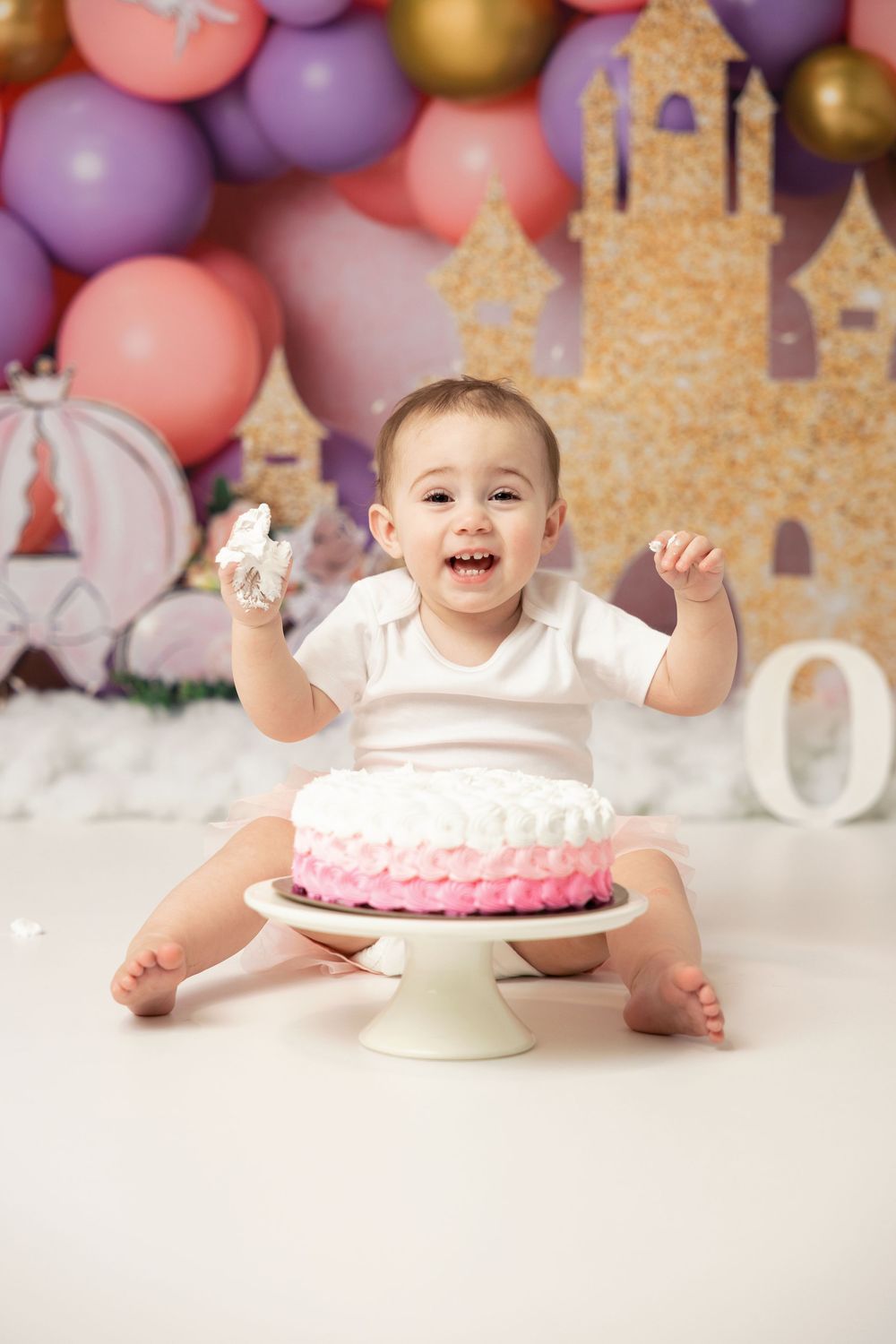 Pink and purple balloon backdrop with cake smash setup featuring a pink ombre cake on white pedestal stand.