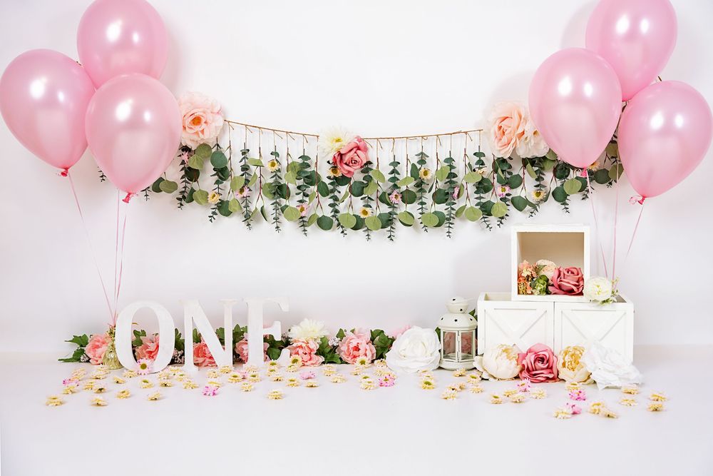 White dessert table decorated with pink balloons, flowers, and 'ONE' letter sign for a first birthday celebration.
