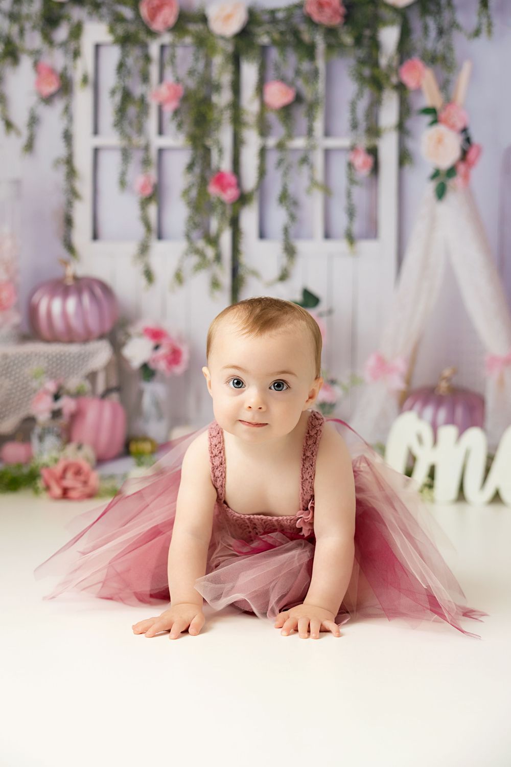 Baby in dusty rose tulle dress crawls on white floor with floral backdrop and pink decorative elements.