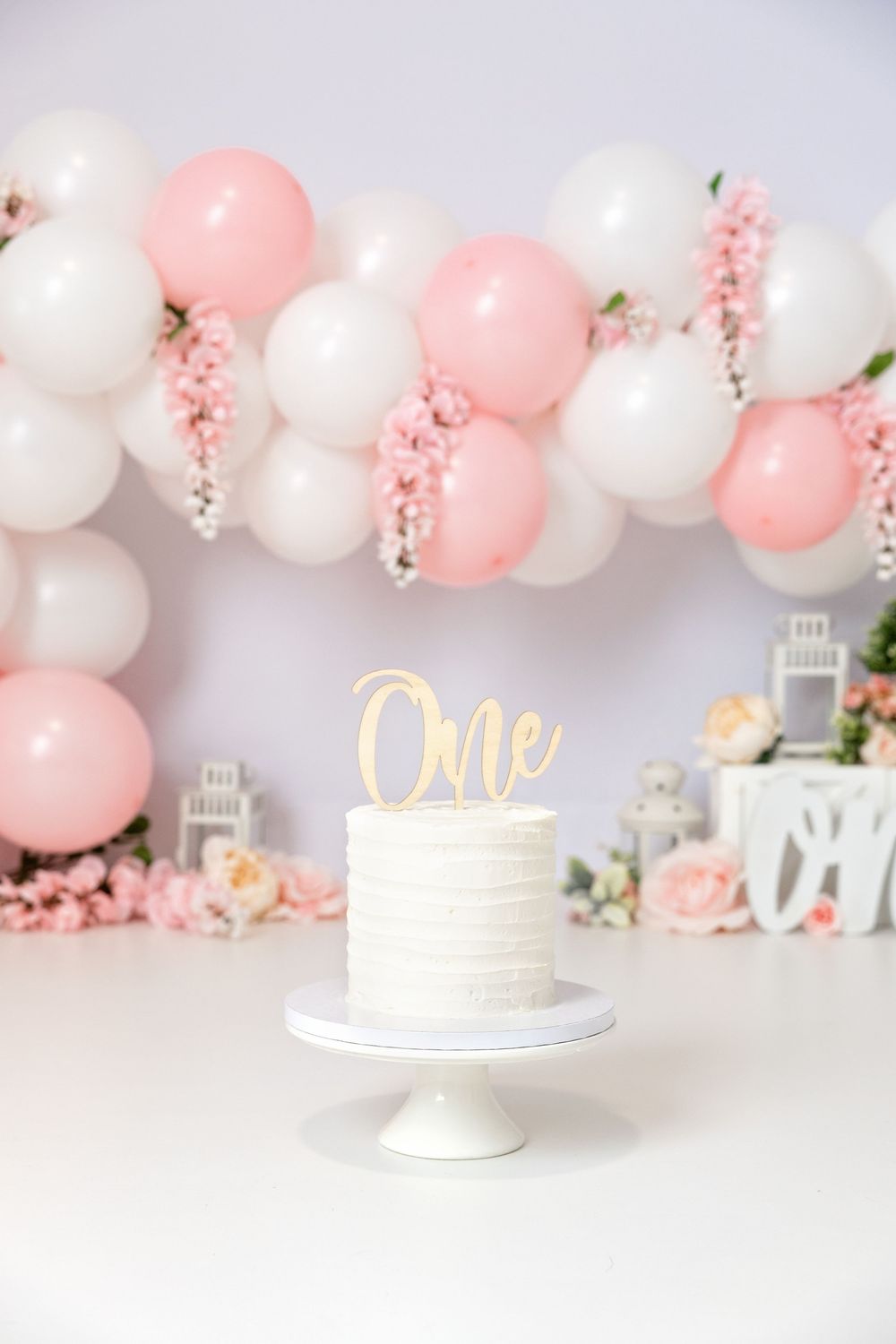 White first birthday smash cake with gold 'One' topper, pink and white balloon garland, floral decor backdrop.