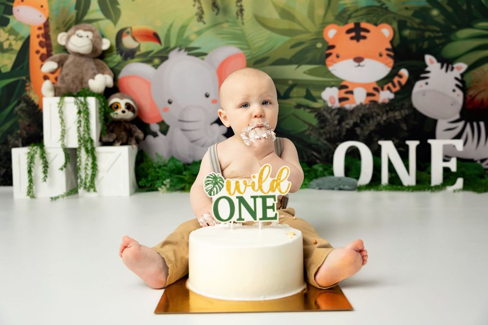Festive first birthday party setup with jungle animal decorations, white cake, and green number blocks against leafy backdrop.