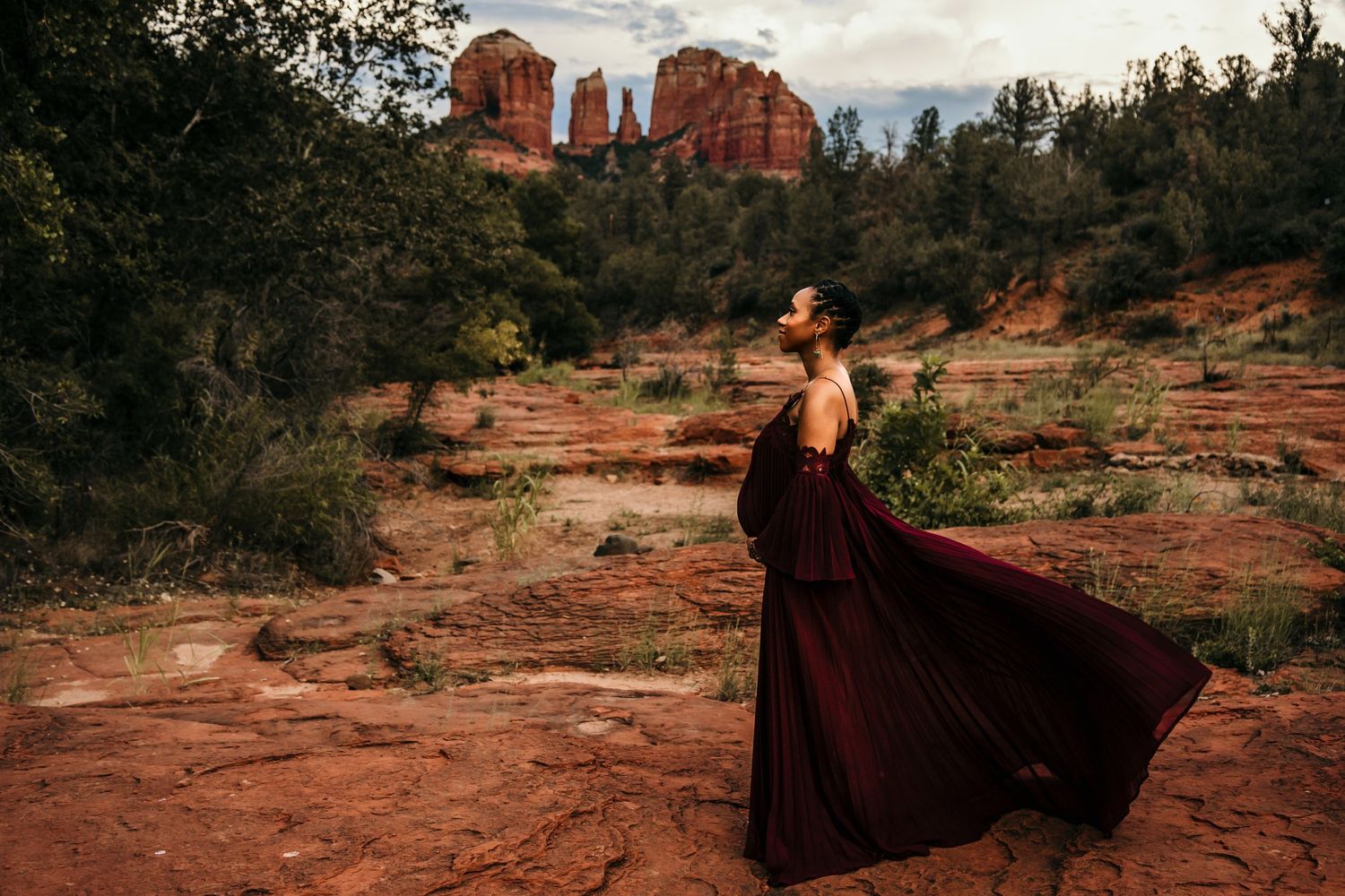 Elegant maternity portrait in burgundy dress with Sedona's red rocks and cloudy sky in background.