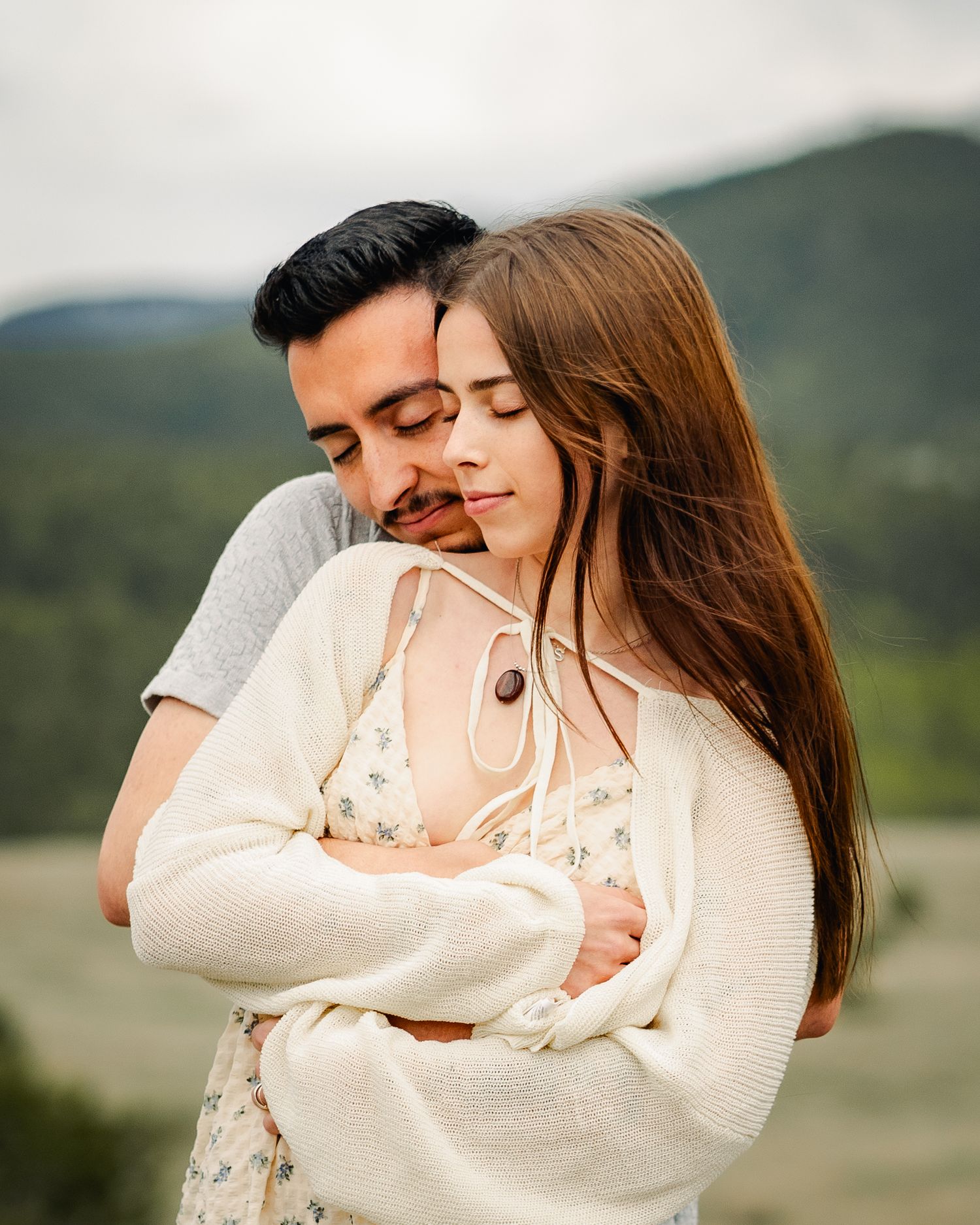 Couple in white sweaters embracing outdoors with mountain scenery in background.