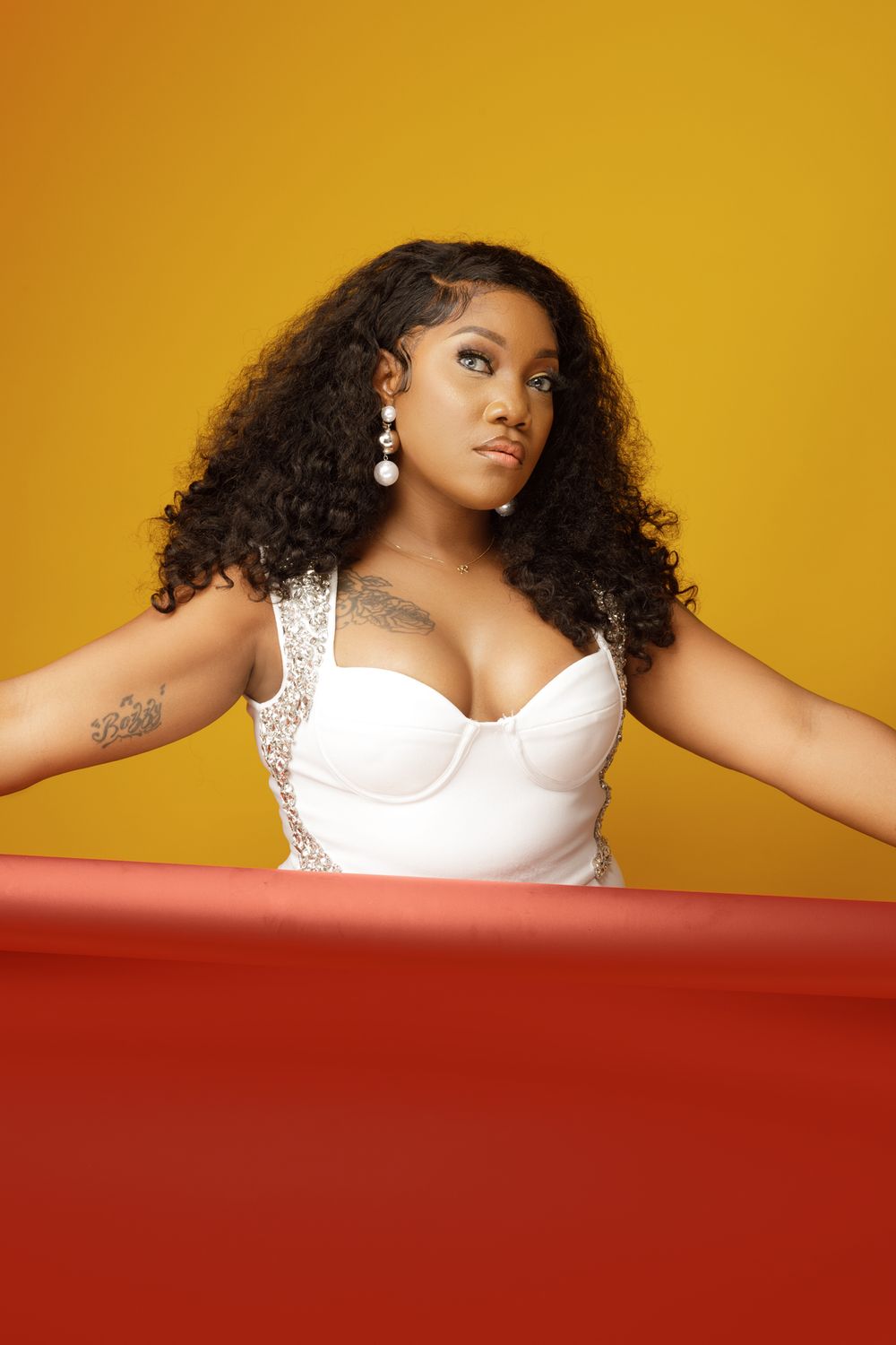 Person with long curly dark hair wearing a white bustier top poses against orange background with red foreground element.
