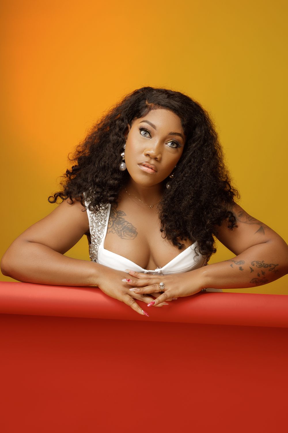 Woman with voluminous curly hair wearing white top poses against orange backdrop with red platform in studio portrait.